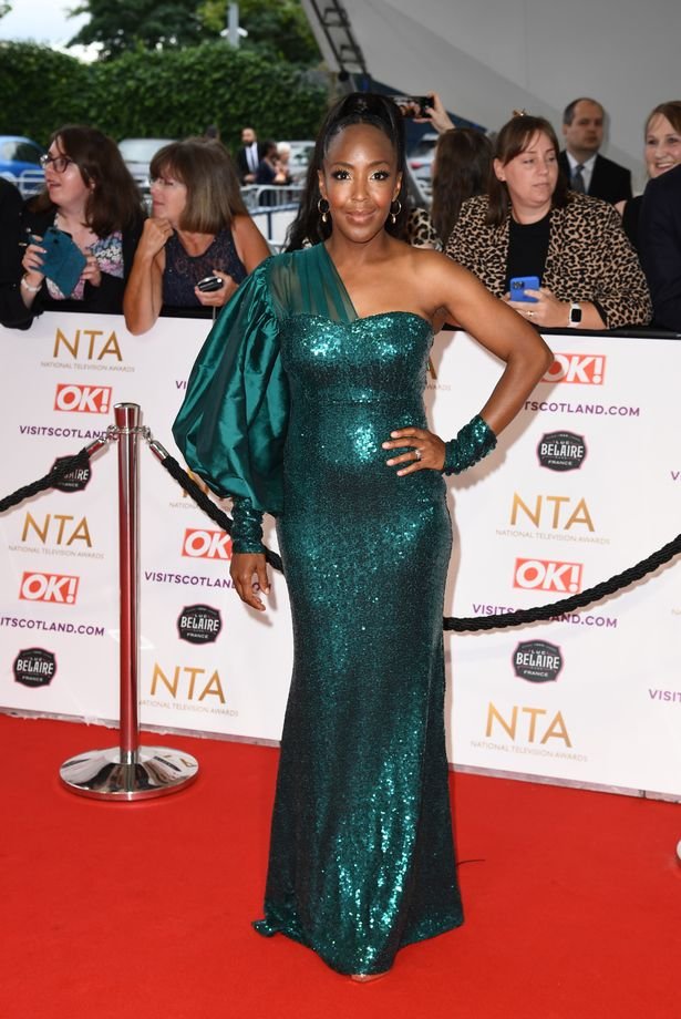 A woman in a sparkly emerald green gown with a matching sleeve on one arm poses on a red carpet at an awards event, with onlookers and a branded backdrop behind her.