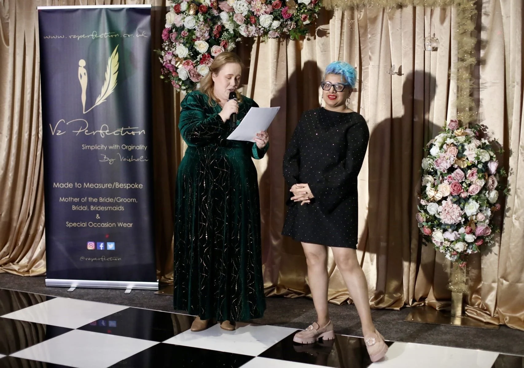 Two women on a stage, one holding a microphone and reading from a paper. Behind them is a large floral arrangement and gold curtains. To the left is a banner with the logo and text for 'Re-Perfection,' a wedding dressmaker, with social media icons at
