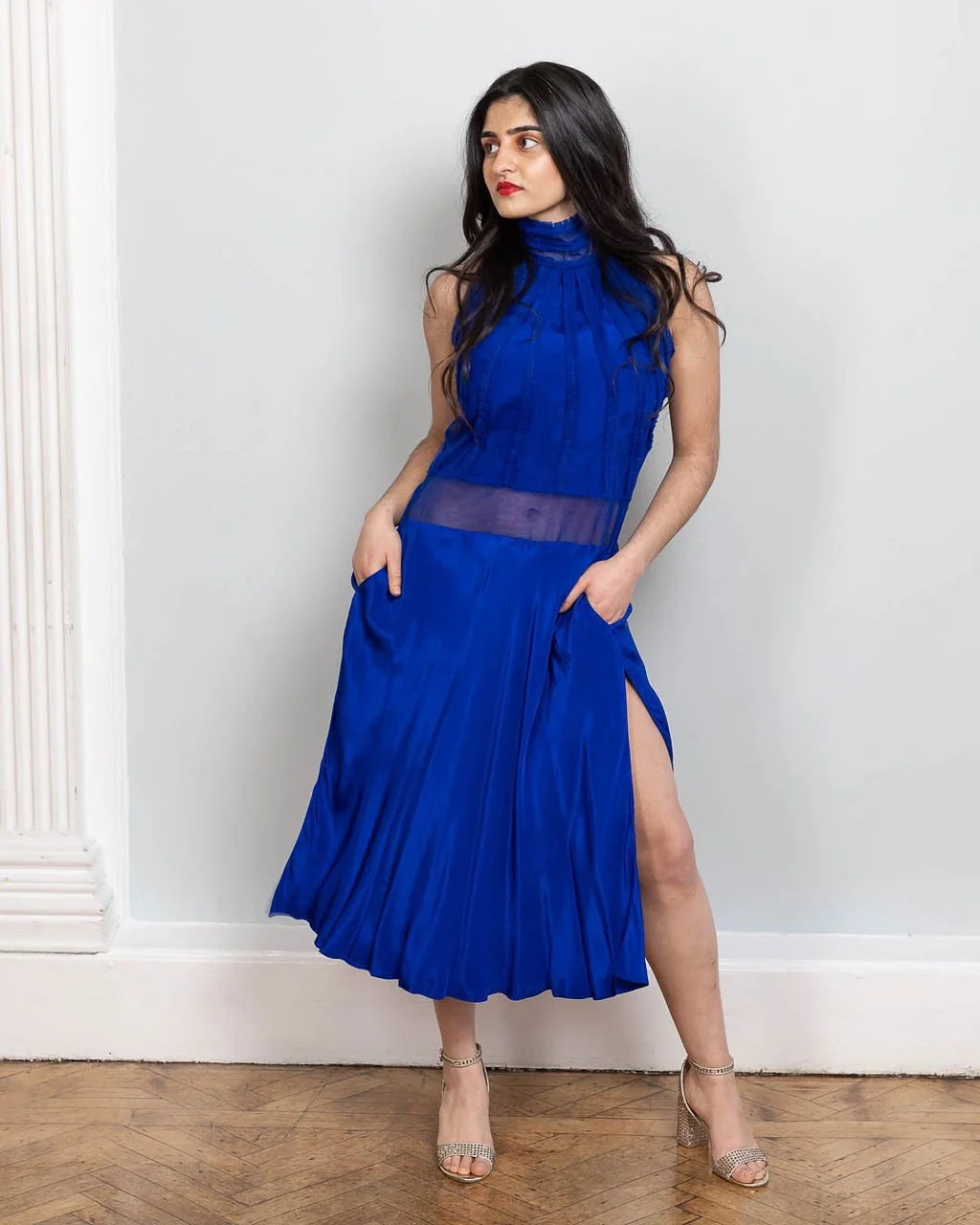 A woman with long dark hair wearing a royal blue dress with a high neck and side slit, standing against a plain white wall with wooden flooring.