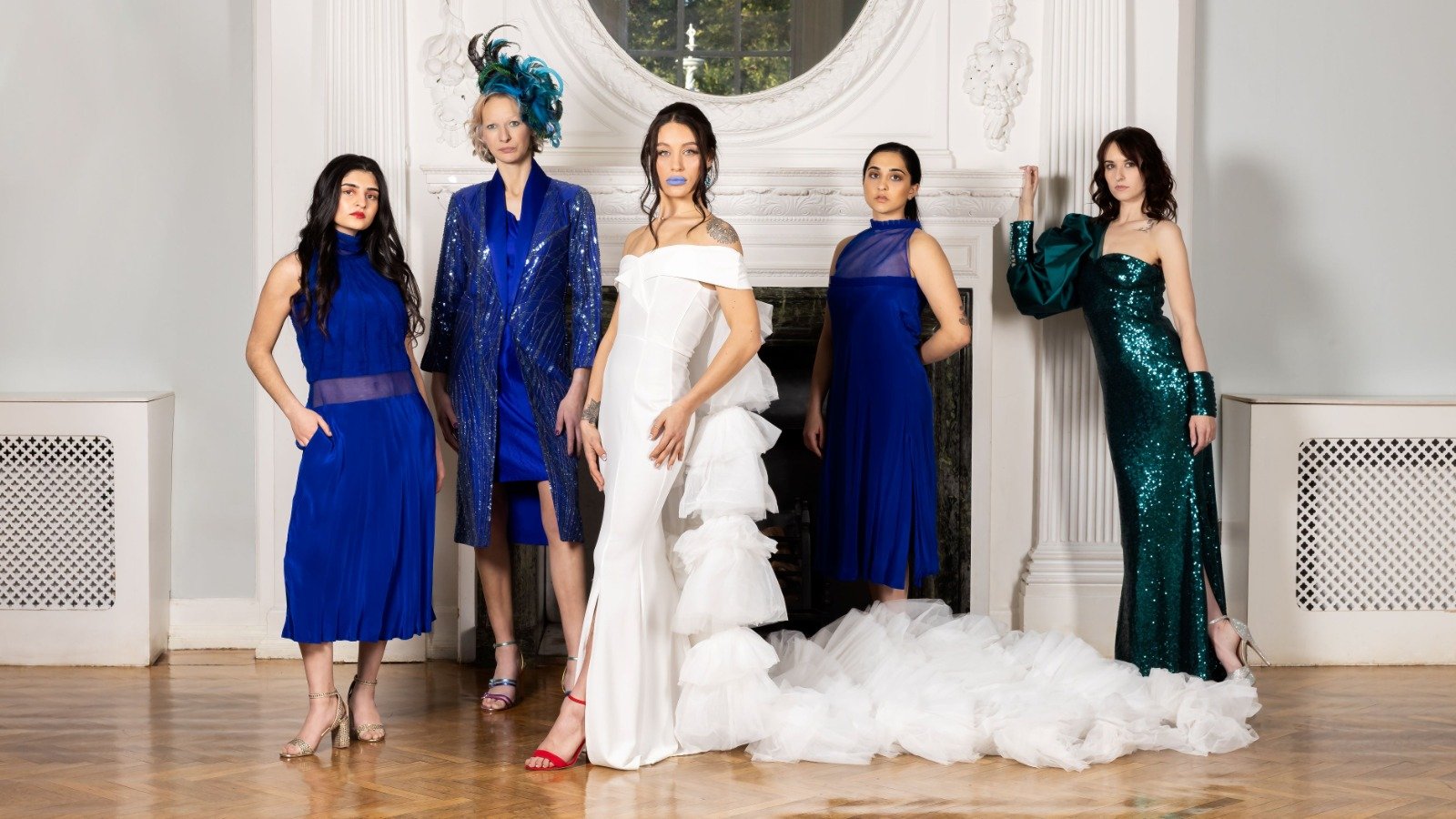 Five women in elegant dresses pose in front of a white fireplace. The woman in the center wears a white wedding gown with ruffles and blue lipstick. The other women wear various shades of blue and green dresses, with different styles and accessories.