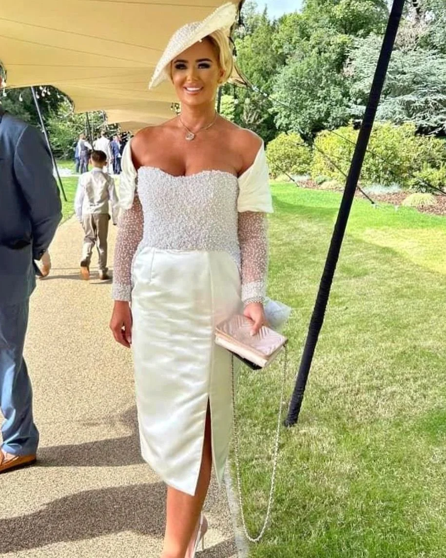 A woman in an elegant white dress with a slit, holding a clutch purse, wearing a large white hat, and standing outdoors on a sunny day at a formal event or wedding, with greenery in the background.