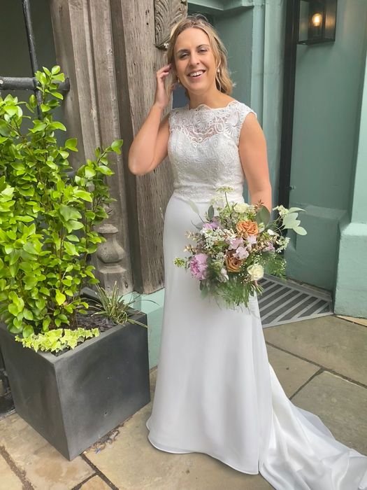 A woman in a white wedding dress holding a large bouquet of flowers, standing outside near a wooden fence and green potted plants, smiling and touching her hair.