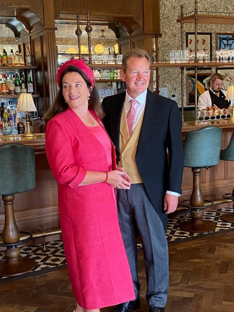 A woman in a pink suit and a man in a dark suit with a yellow vest and pink tie standing together at a bar in a vintage-style room, with a bartender preparing drinks in the background.