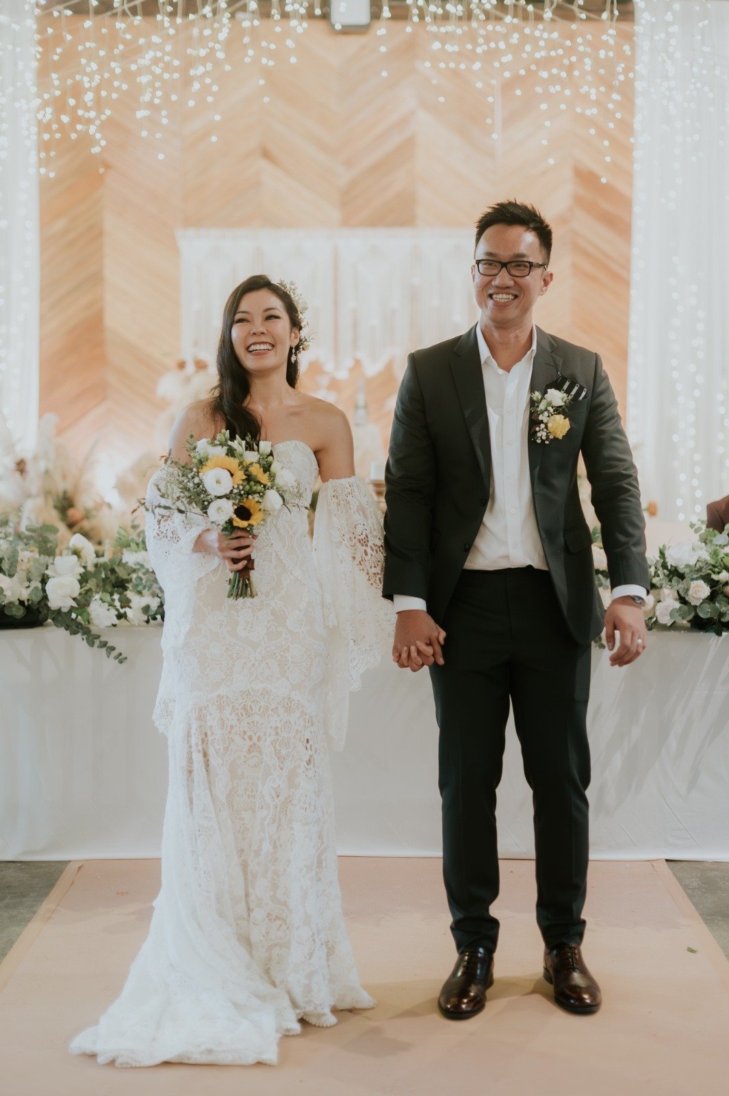 A bride and groom holding hands and smiling at their wedding ceremony, with floral decorations and string lights in the background.