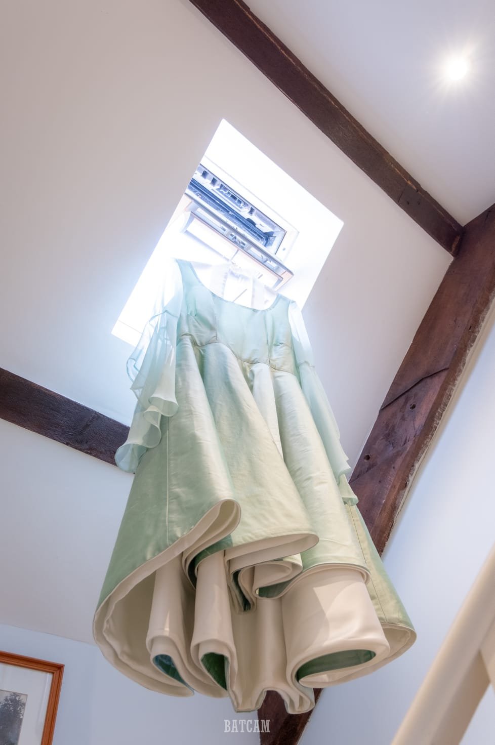 A wedding dress with puffy sleeves hanging in front of an open window in a white-walled room with wooden beams.