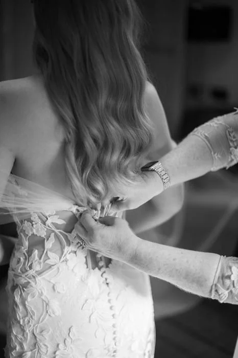A woman is getting assisted with her wedding dress by another person, likely helping her fasten the back of her dress.