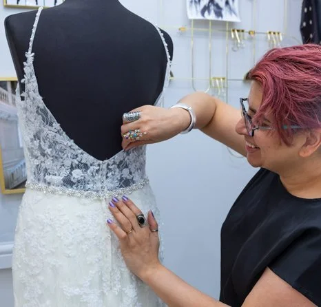 A woman with pink hair and glasses adjusting a white lace wedding dress on a mannequin.