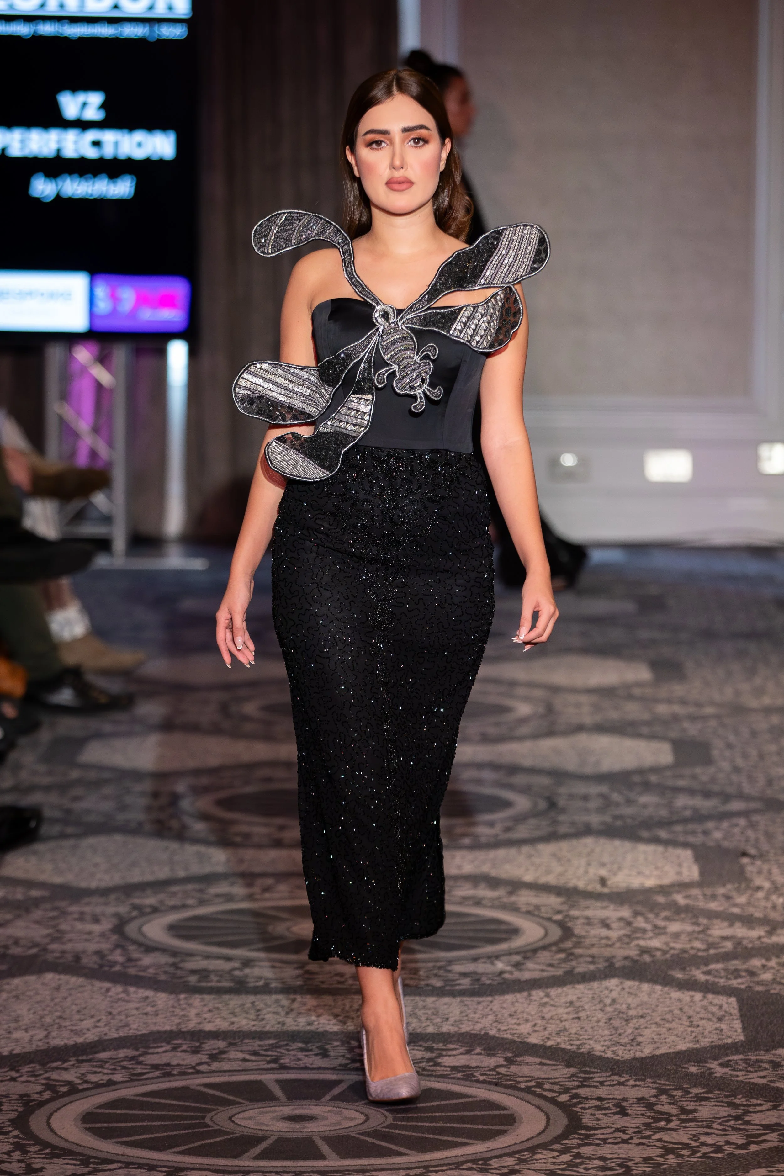A woman walking on a runway at a fashion show, wearing a black, form-fitting, sequined dress with a butterfly-shaped accessory on her chest. She has shoulder-length brown hair and neutral makeup.
