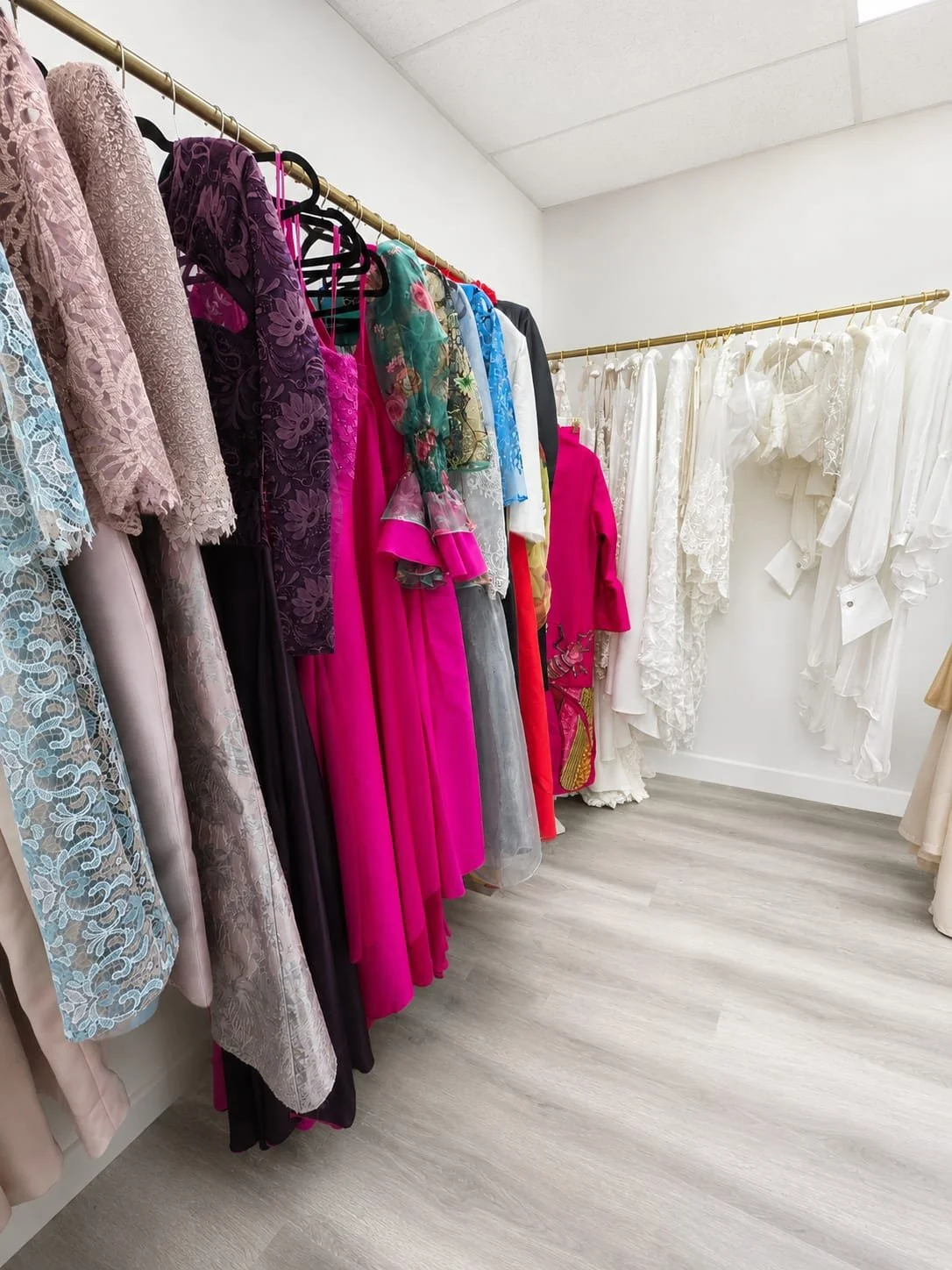 A boutique or dress shop with racks of colorful dresses on the left and mostly white dresses on the right, hanging on golden bars in a room with white walls and wood-like flooring.