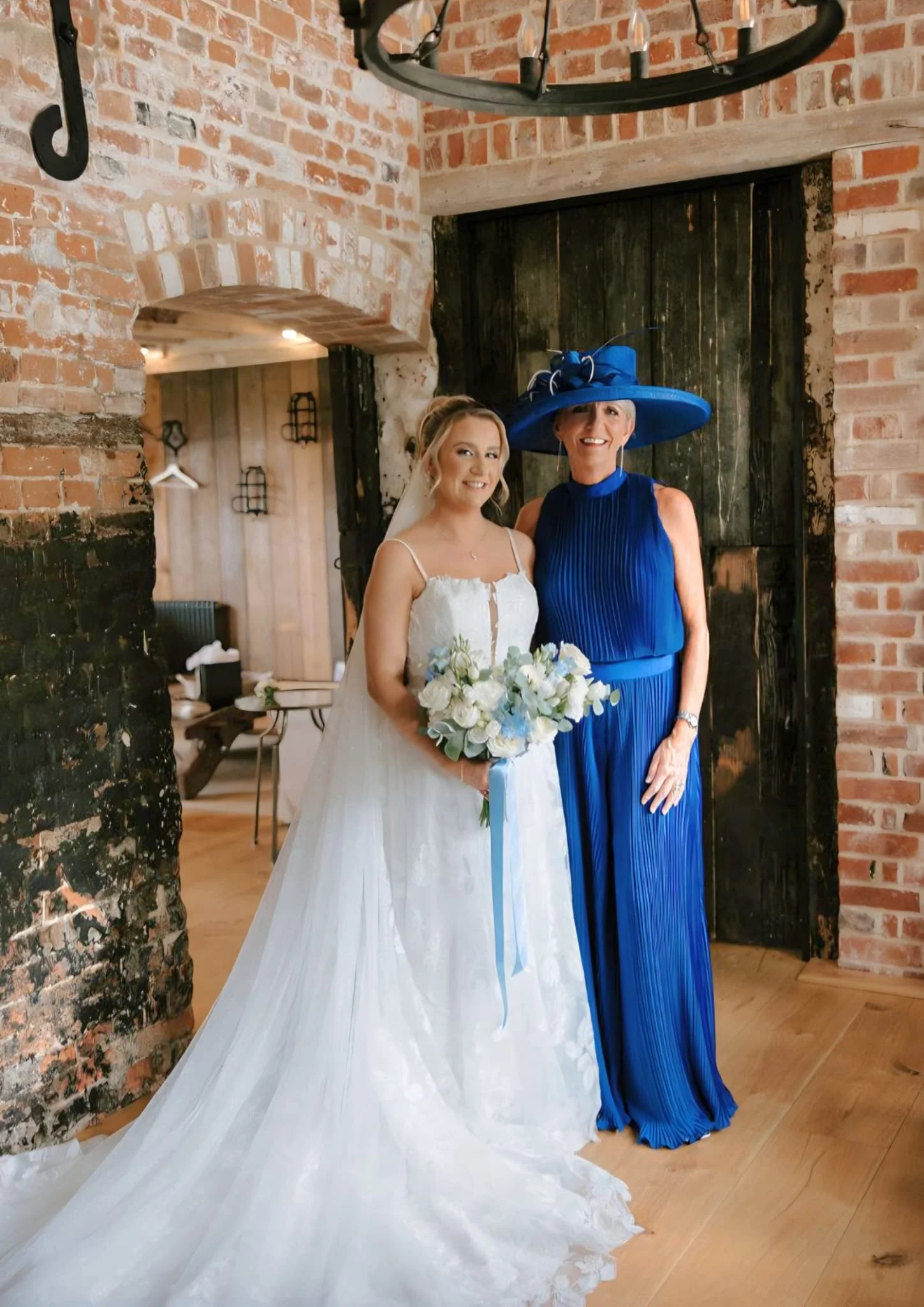 Two women at a wedding, one in a white wedding dress holding a bouquet of white flowers, the other in a royal blue dress and large matching hat. They are standing inside a rustic brick building with black wood doors and warm lighting.