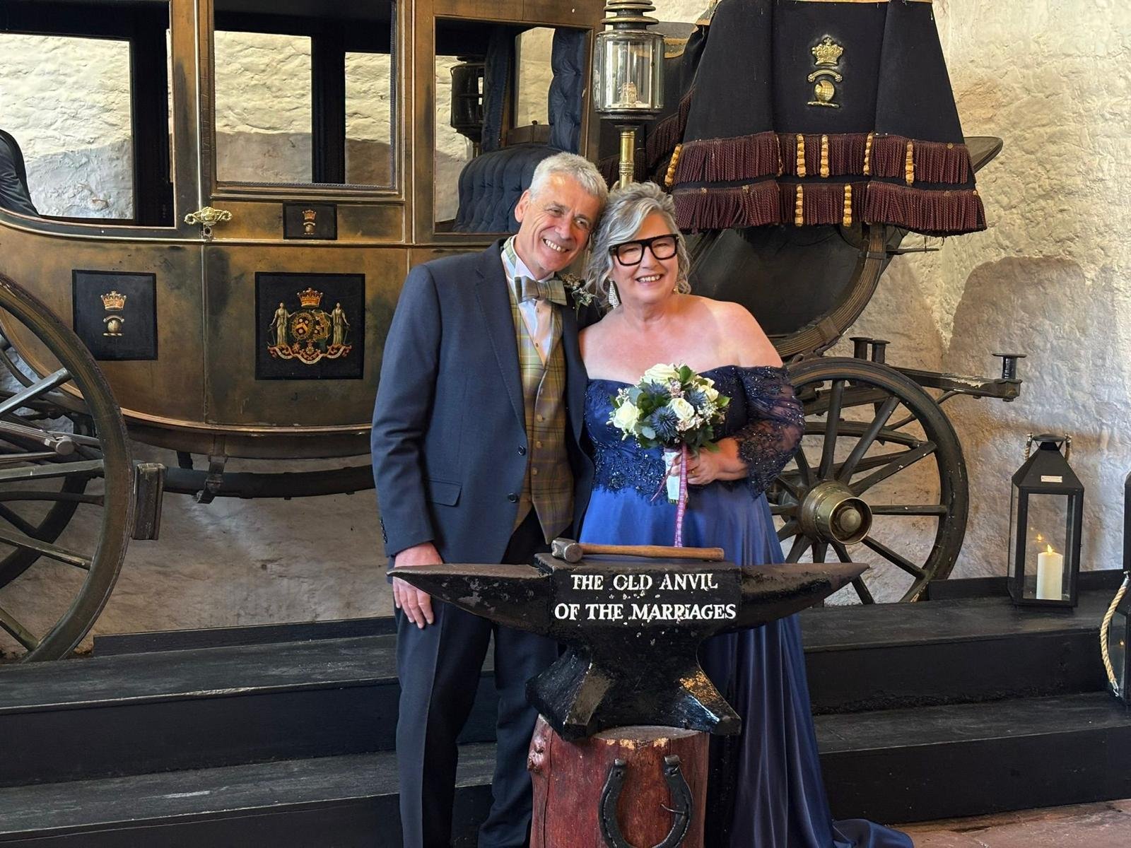 A smiling couple dressed in formal wedding attire standing in front of a vintage carriage and an anvil with the sign "The Old Anvil of the Marriages." The woman is holding a bouquet of flowers, and they are inside a rustic setting with a lantern and 