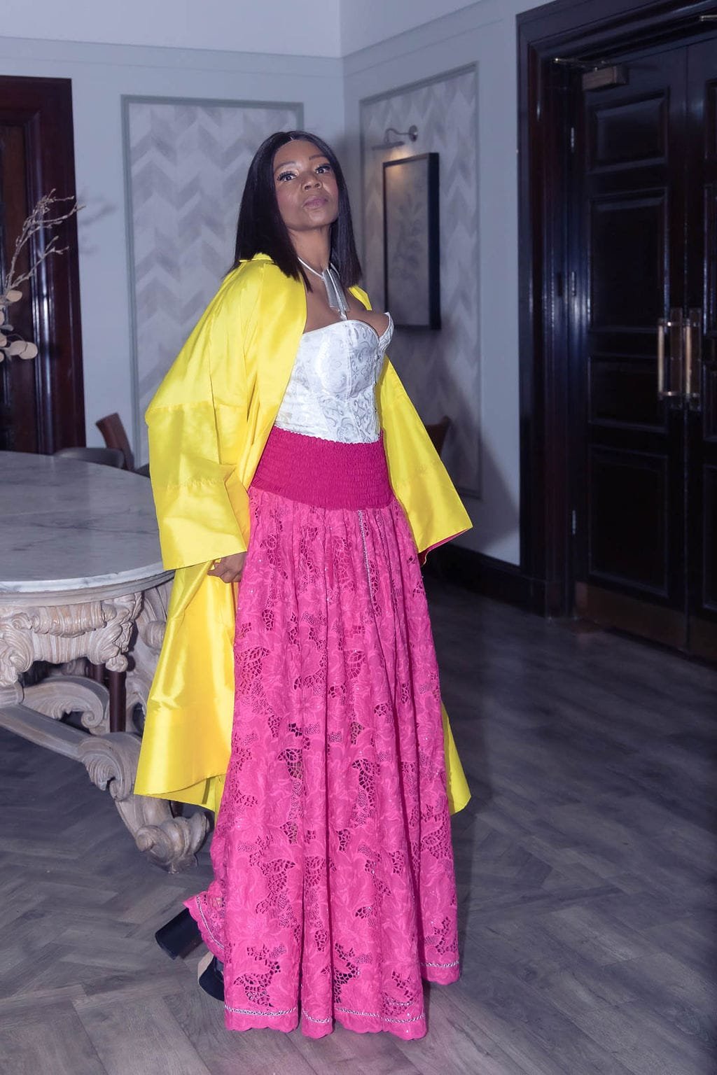 A woman standing in a room, wearing a white strapless top, a vivid pink lace skirt, and a yellow coat draped over her shoulders, with dark hair and makeup.