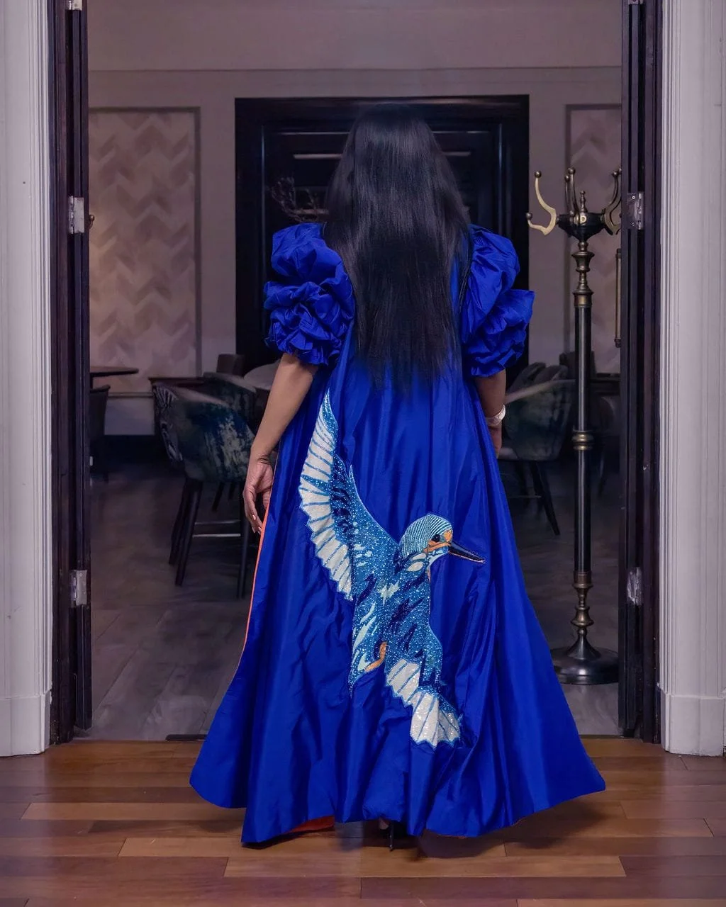 A woman with long dark hair wearing a vibrant blue gown with an embroidered bird design on the back, standing in a doorway facing away.
