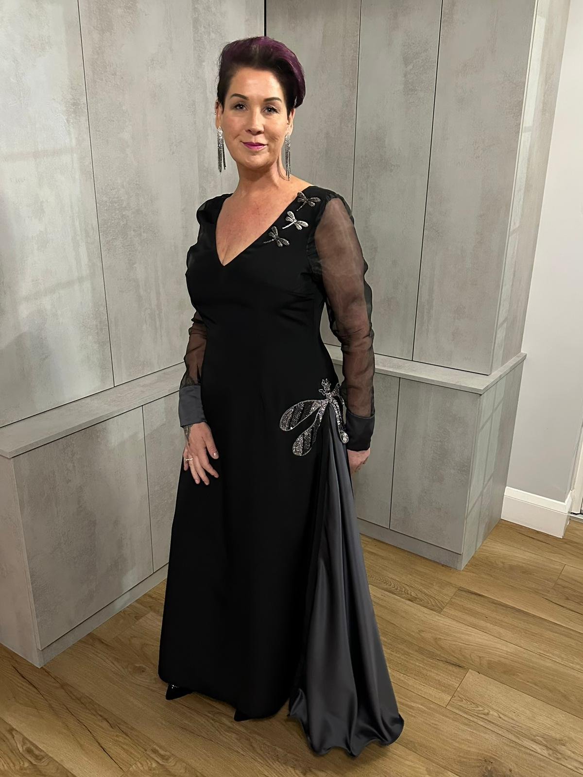 A woman in a black gown with sheer sleeves and dragonfly embellishments poses indoors against a gray wall.