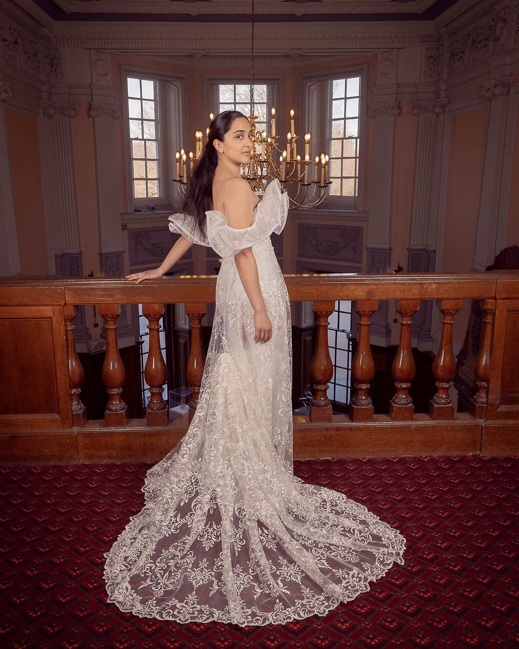 Woman in an elegant lace wedding gown with puffy sleeves, standing on a balcony inside a grand, historic building with ornate moldings, tall windows, and a large chandelier.