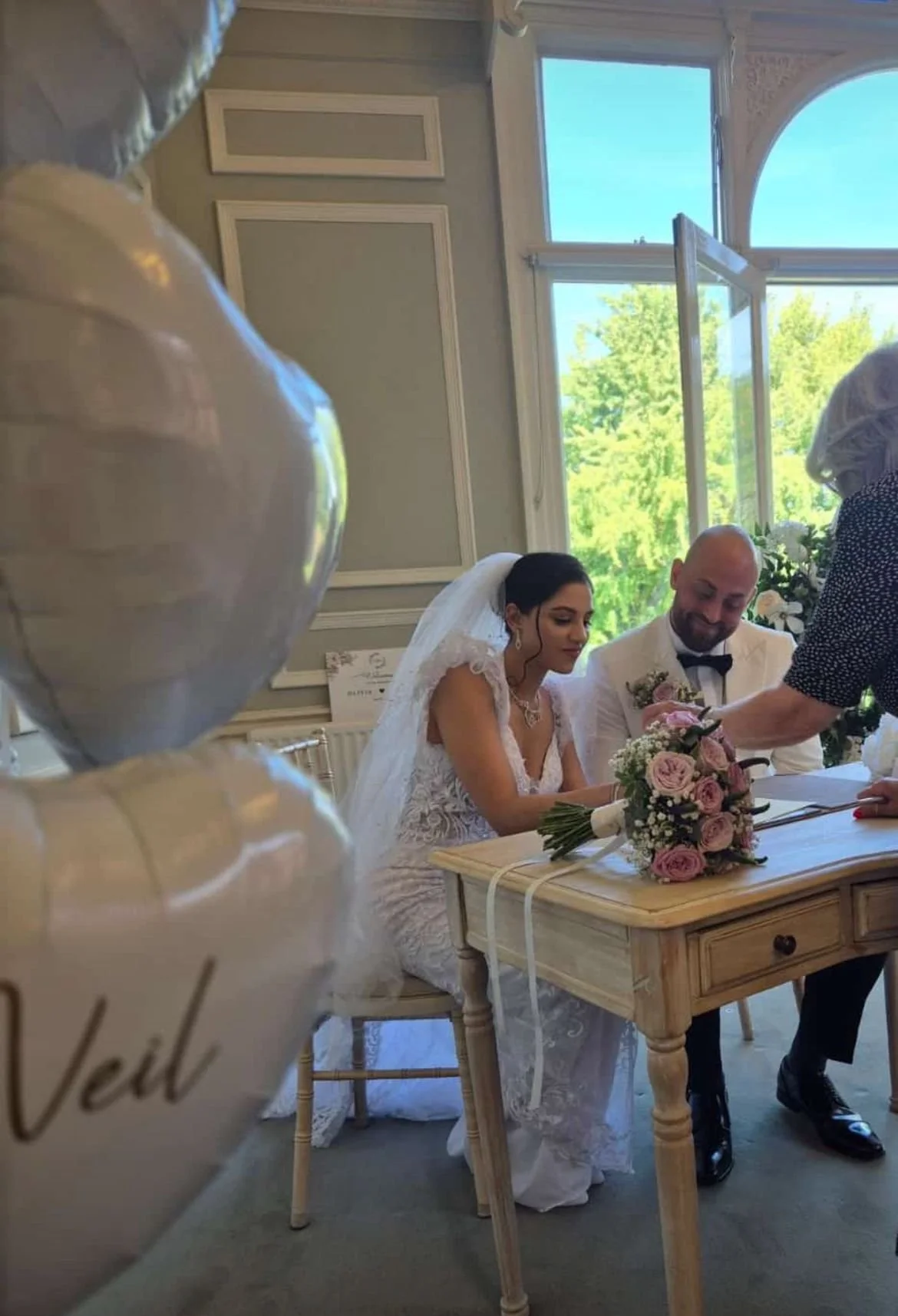 A bride and groom seated at a small wooden table during their wedding ceremony, with a woman helping them sign documents. The bride is wearing a wedding dress and veil, and the groom is in a tuxedo. A large bouquet of pink roses and other flowers is 