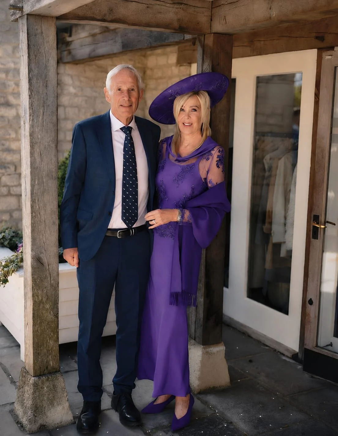 An elderly man in a blue suit and a white shirt with a blue tie, standing next to a woman in a purple dress and matching hat, both smiling and posing together outside near a wooden door.