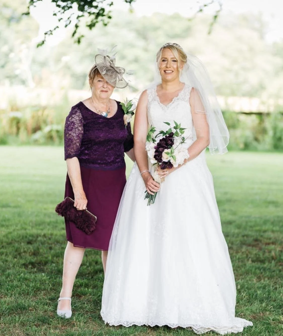 A bride and an older woman standing outdoors on grass, both dressed elegantly. The bride wears a white gown, lace details, a veil, and holds a bouquet. The older woman wears a dark purple lace top, a matching skirt, a large hat with feathers, and hol