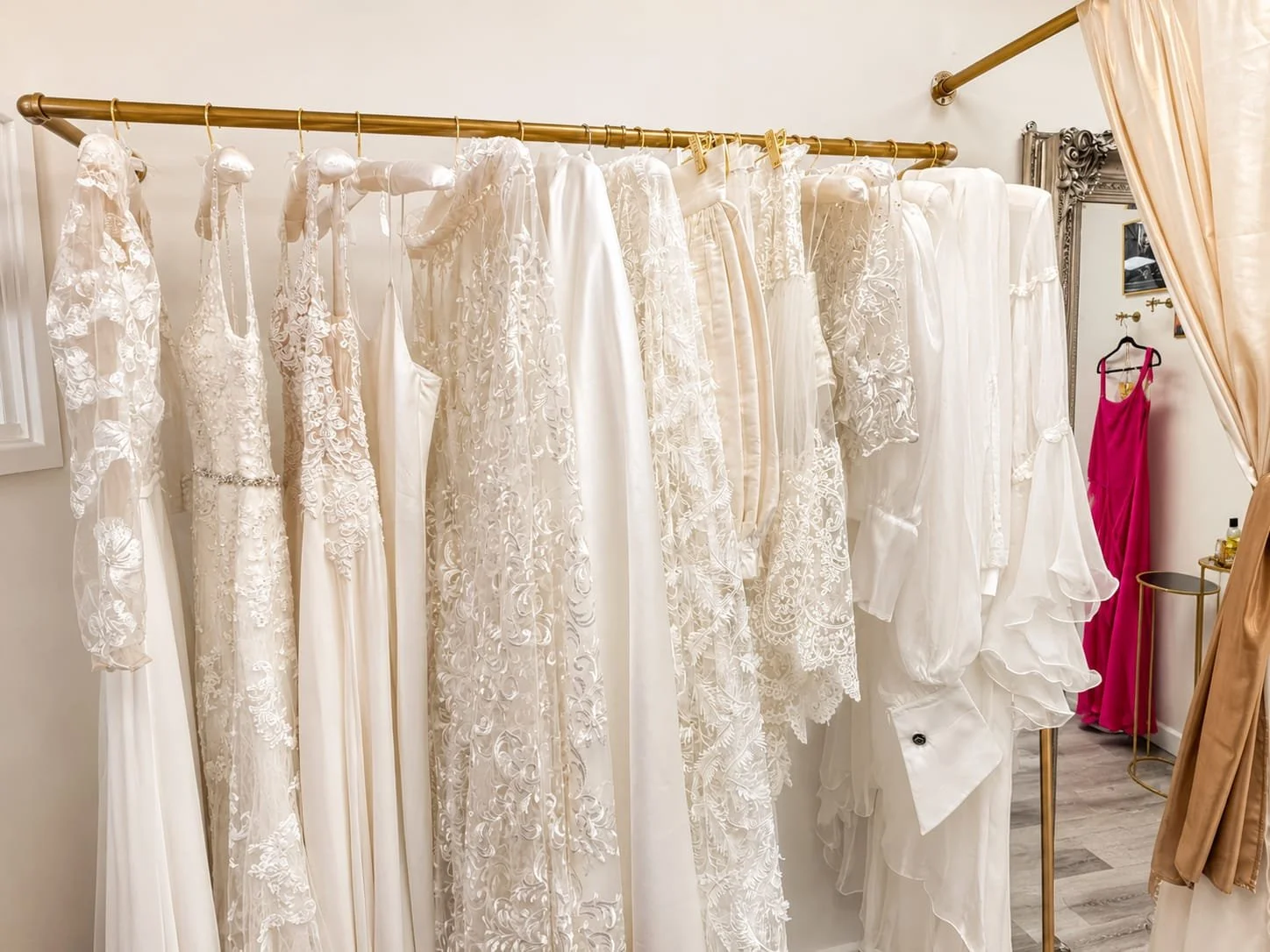A clothing rack with various white wedding dresses with lace and embroidery details, and a pink dress hanging in the background.
