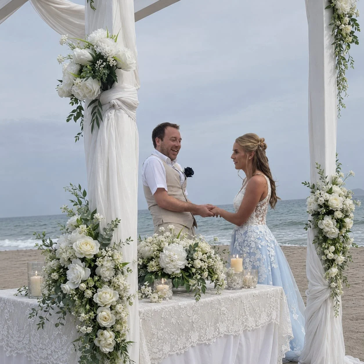 A bride and groom holding hands and smiling during a beach wedding ceremony under a floral arch with white flowers and candles.