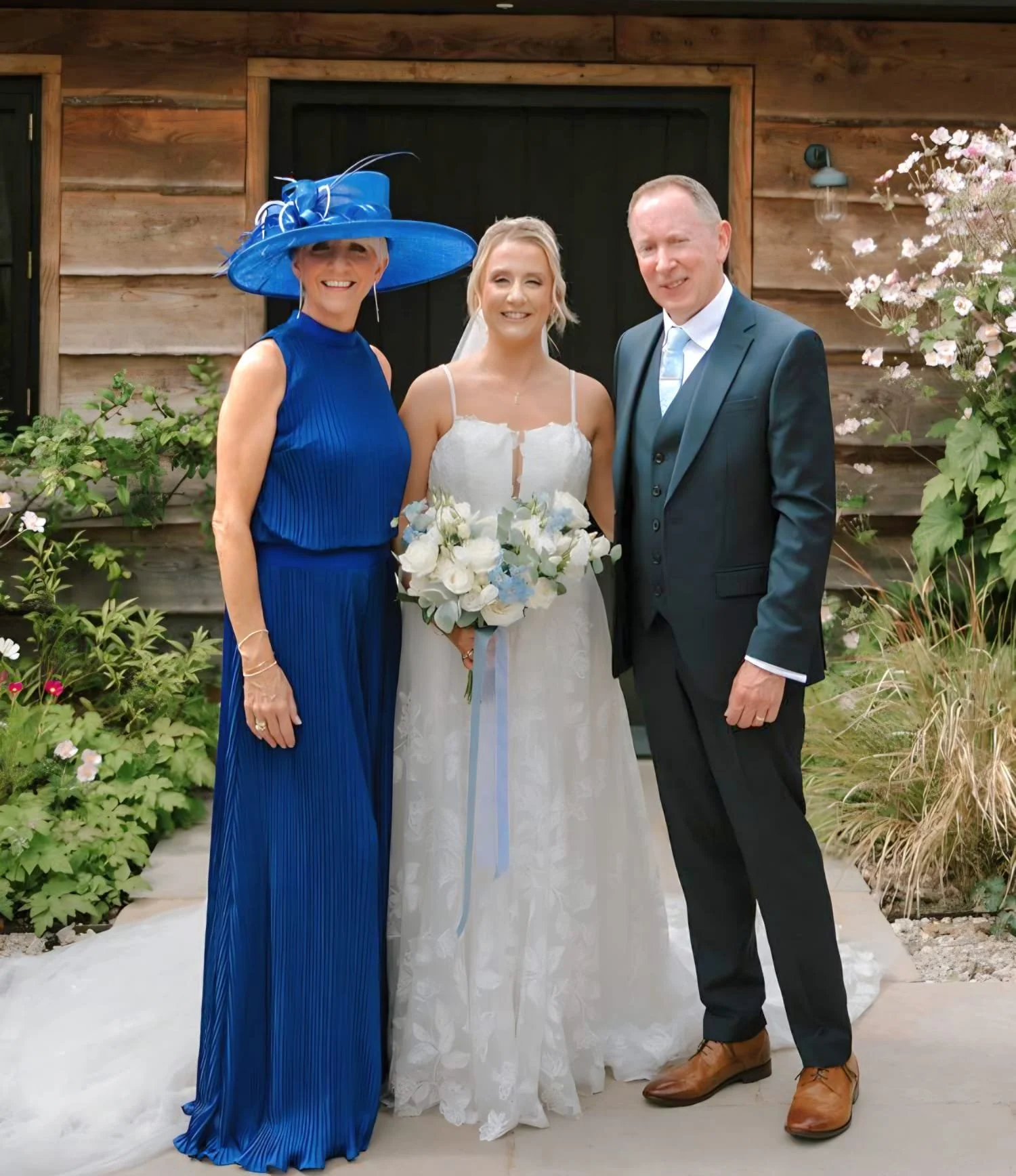 A woman in a white wedding dress holding a bouquet of white flowers, standing between a woman in a blue dress with a large matching blue hat, and a man in a dark suit with a tie, in front of a rustic wooden building with plants and flowers surroundin