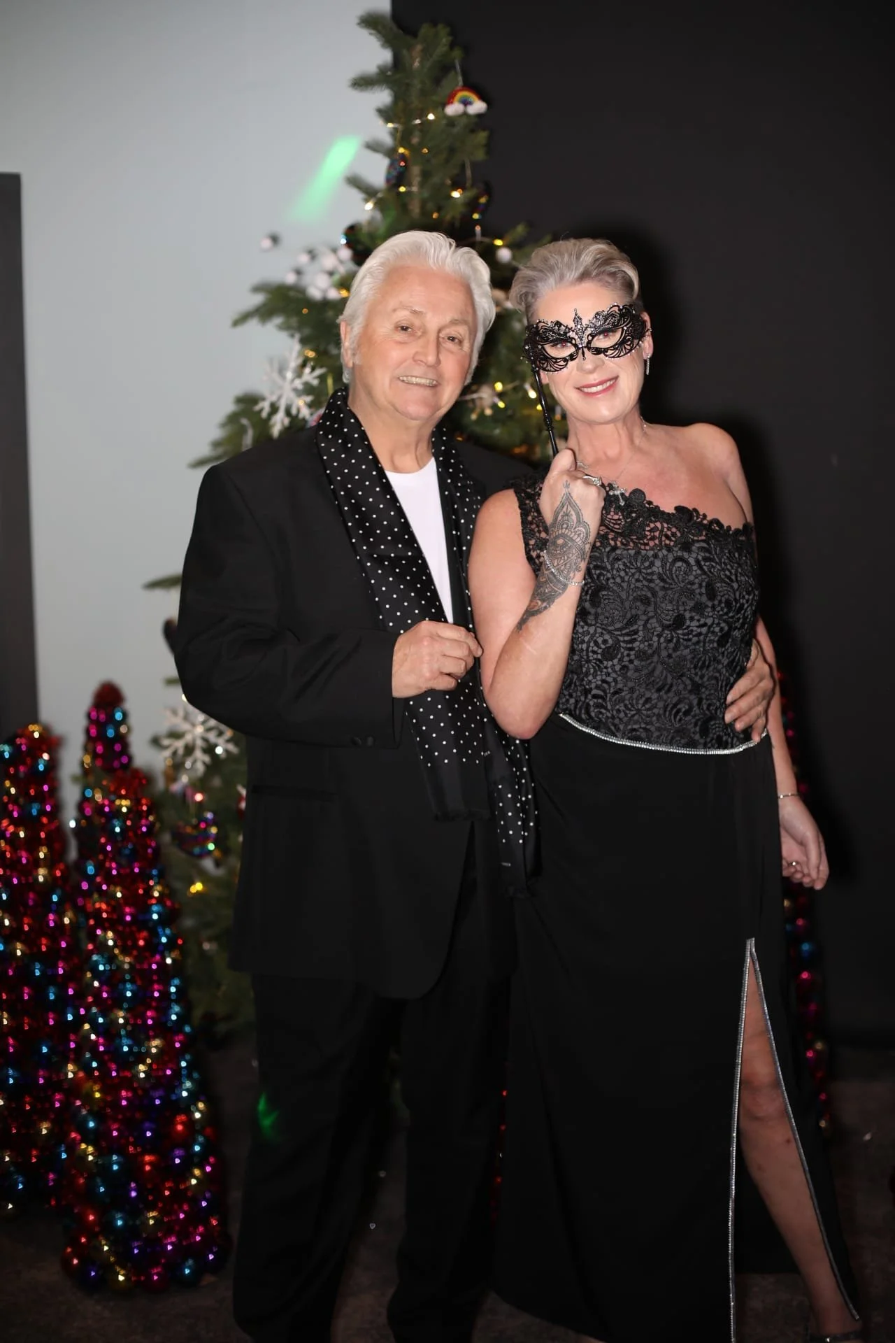 An elderly man and woman standing in front of a decorated Christmas tree. The woman is holding a black lace mask and is dressed in an elegant black dress. The man is wearing a black suit with a polka dot scarf.