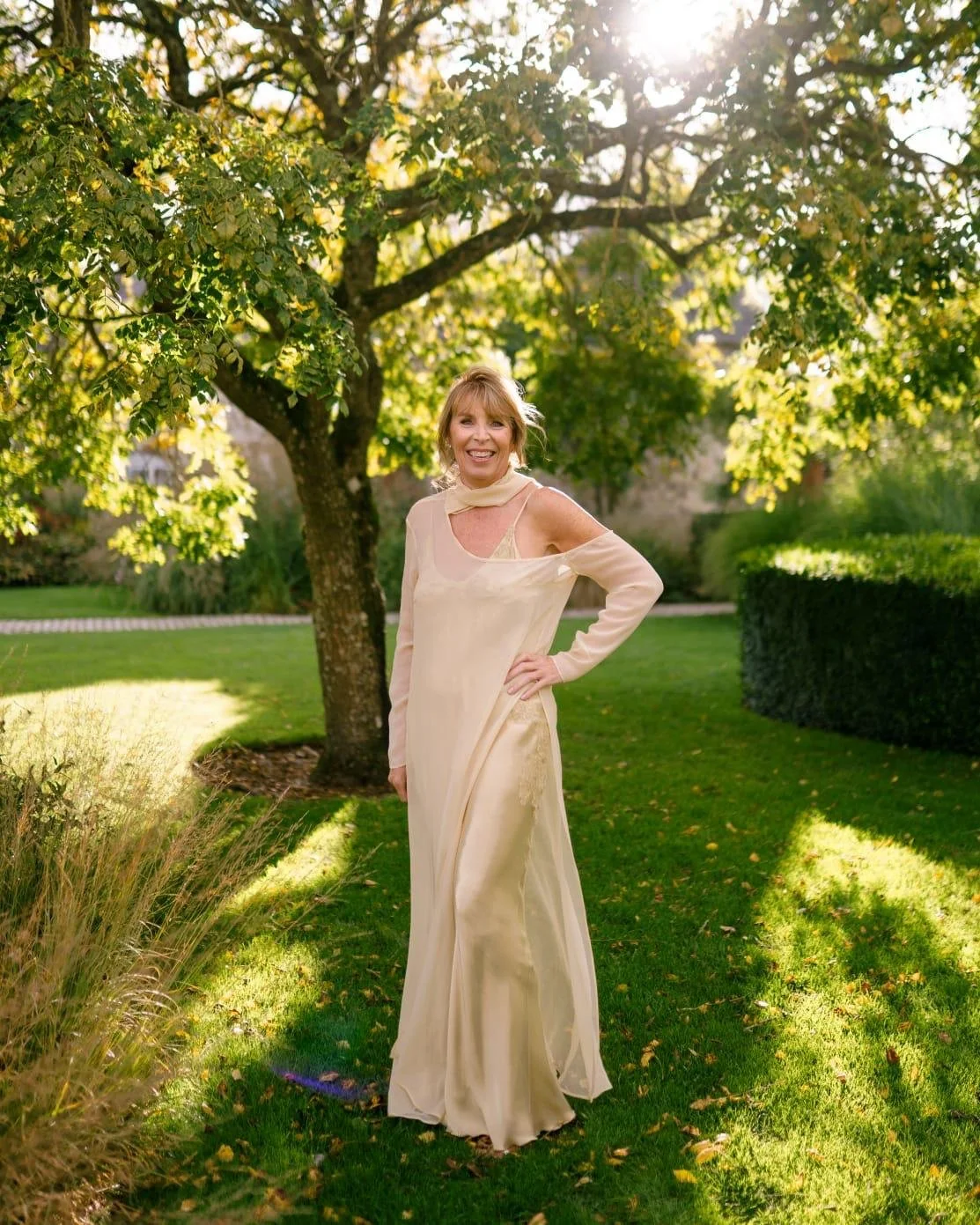 A woman standing outdoors on a sunny day, wearing a long cream-colored dress, with a tree and lush green lawn in the background.