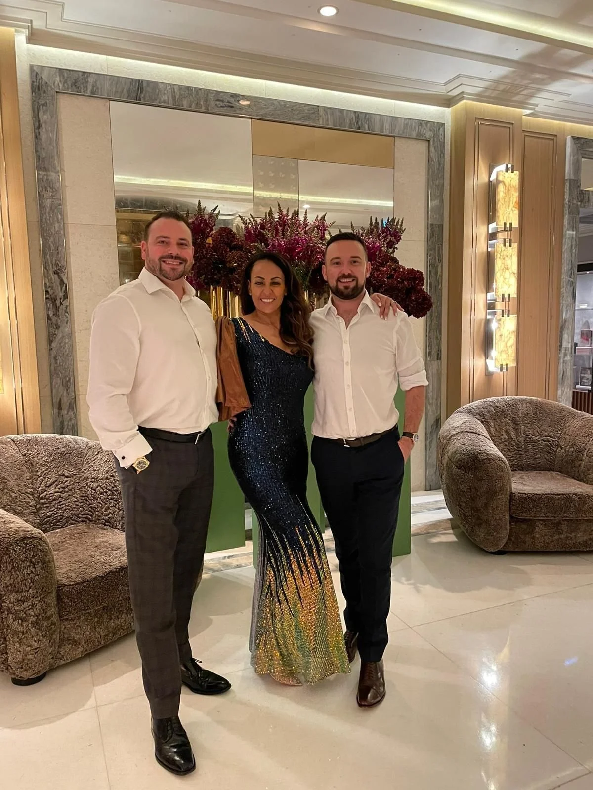 Three people standing together in a hotel lobby, smiling for the photo. The woman in the middle is wearing a sparkling, colorful evening gown, while the two men on either side are dressed in white shirts and dark pants. Behind them is a large floral 