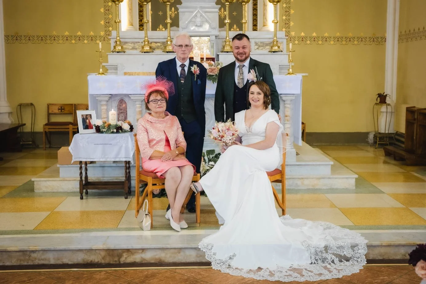 A wedding ceremony inside a church with five people. The bride is seated in a white dress holding a bouquet, with an older woman in pink sitting next to her. Two men in suits stand behind them. The altar is decorated with candles, flowers, and a fram