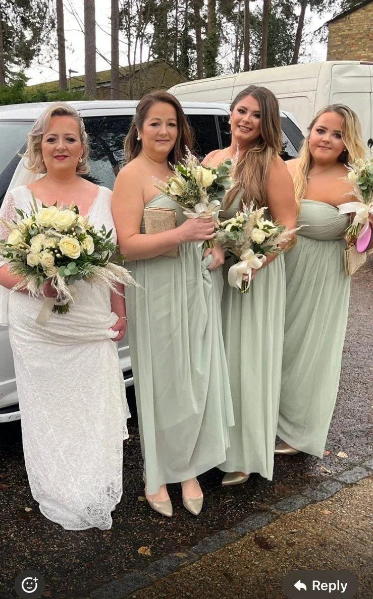 Four women dressed in elegant light-colored dresses holding bouquets of flowers, standing outdoors near parked vehicles on an overcast day.
