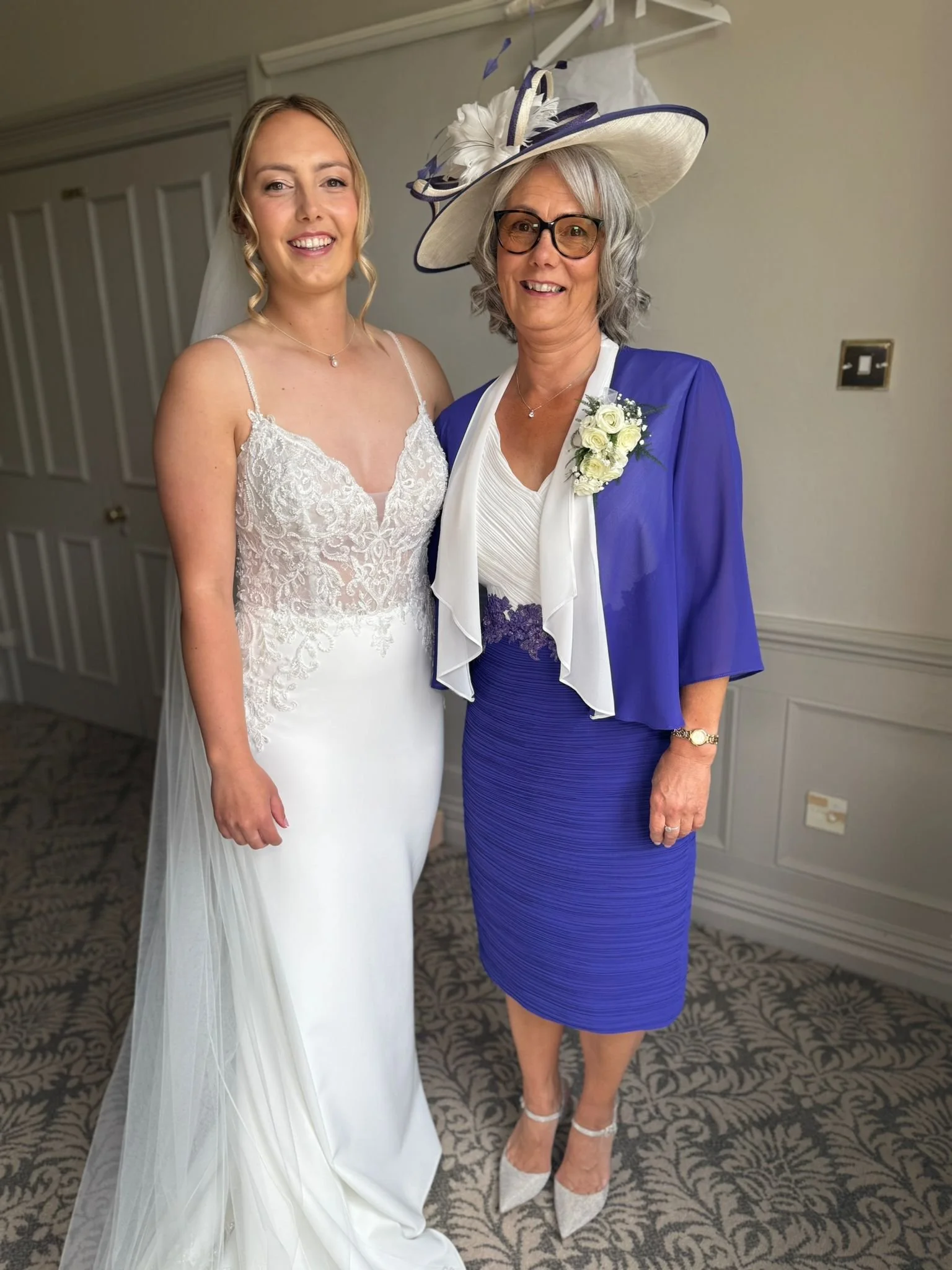 A bride in a white wedding dress standing next to an older woman in a blue dress with a large hat and glasses. The woman has a white flower corsage, and they are smiling inside a decorated room.