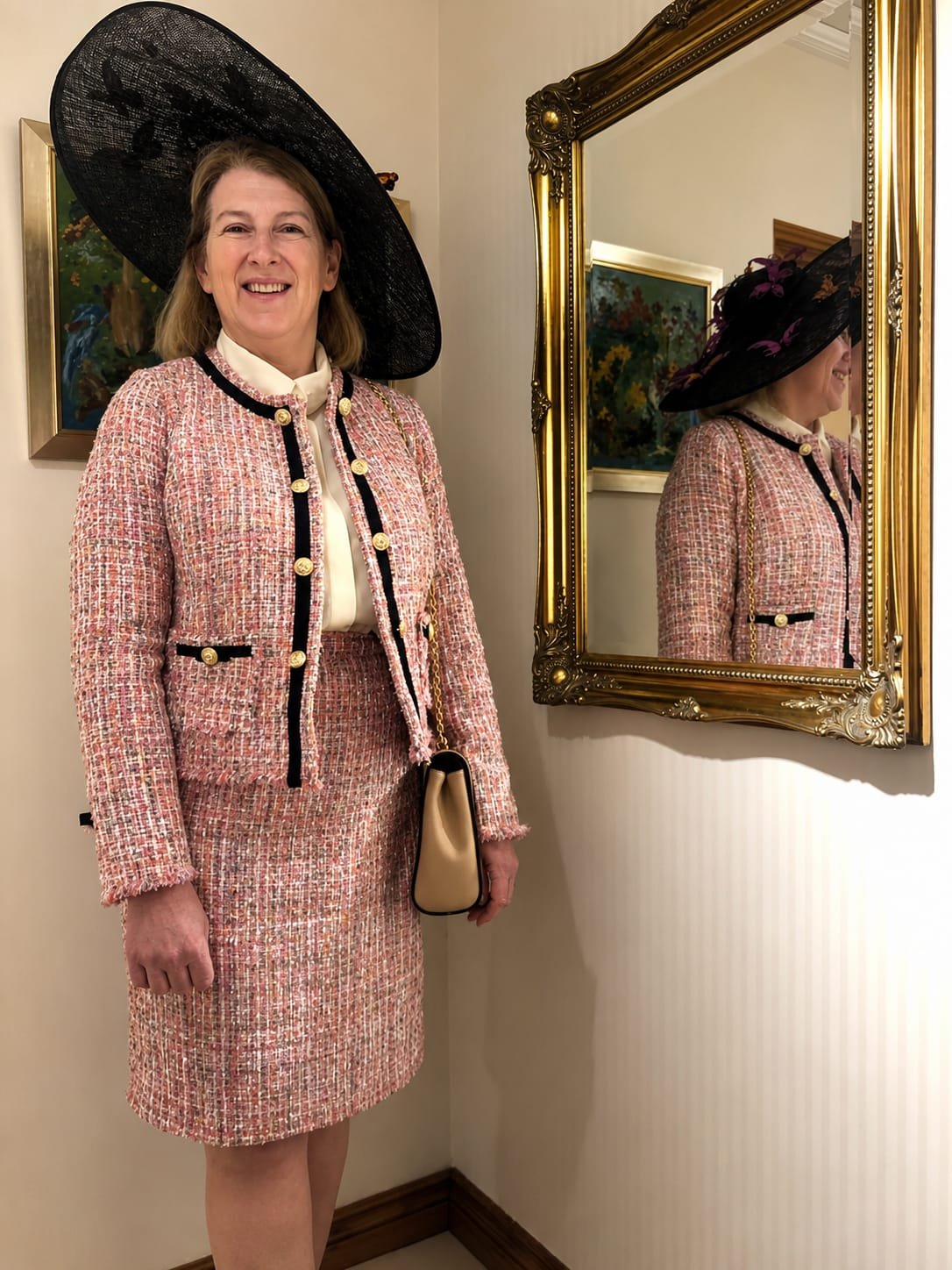 A woman wearing a pink tweed skirt and jacket, with a white blouse underneath, accessorized with a large black hat with purple accents, posing near a gold-framed mirror that reflects her profile, in an interior setting with paintings on the wall.