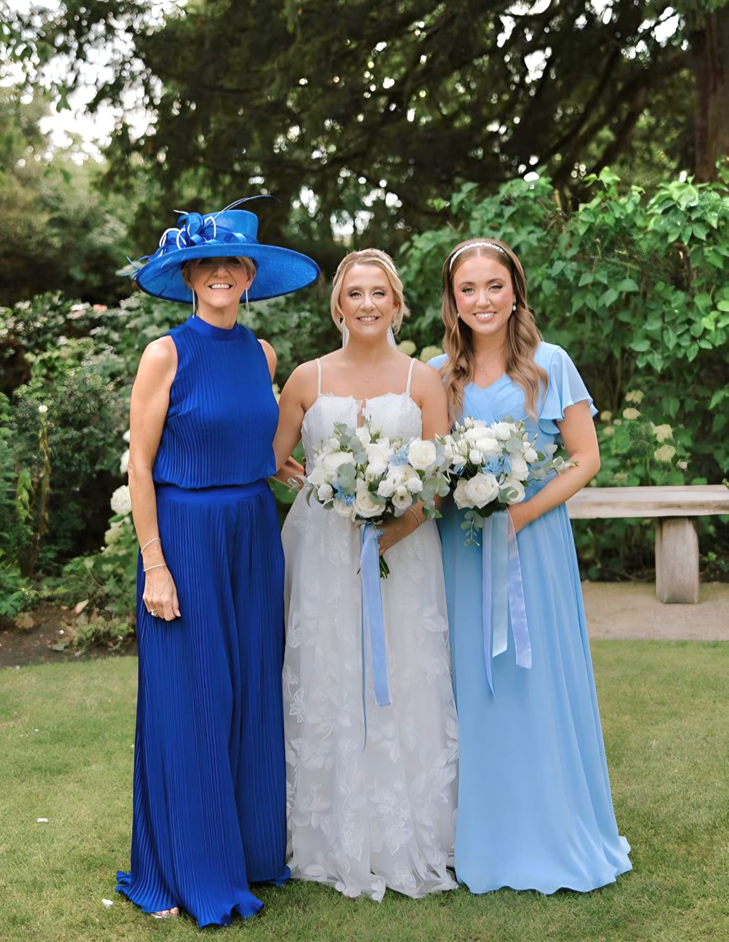 Three women standing outdoors in front of green foliage, dressed in formal dresses. The woman in the center is wearing a white wedding dress and holding a bouquet of white and blue flowers. The woman on the right is wearing a light blue dress, and th
