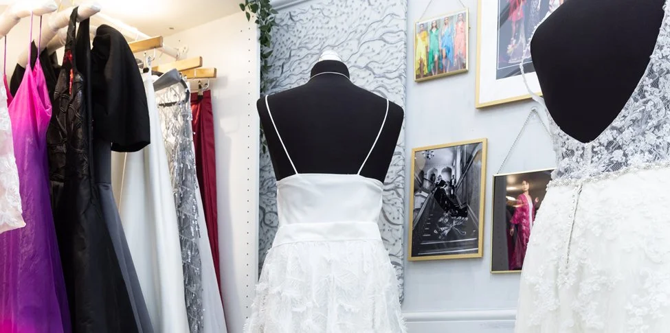 Display of various dresses including bridal gowns, on mannequins and hangers in a boutique or dress shop.