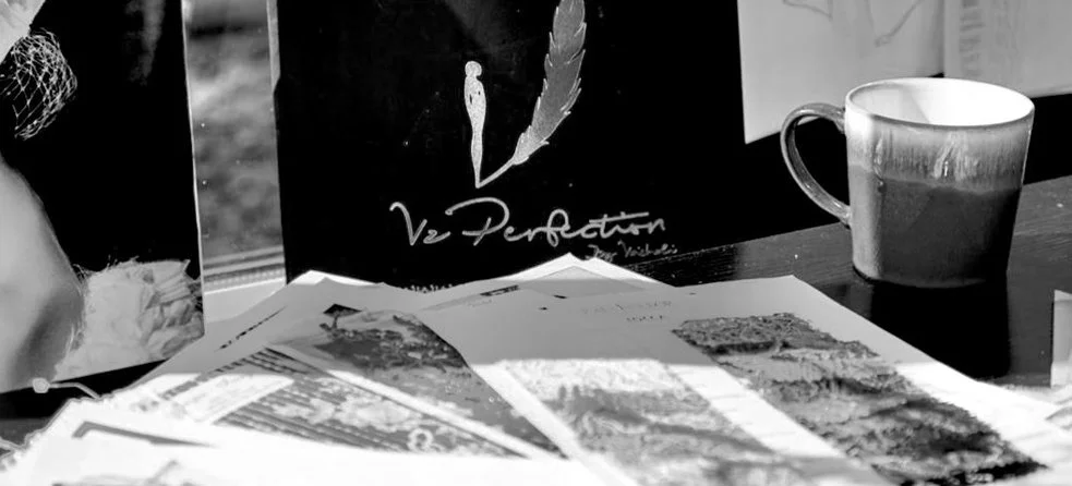 A table with newspapers, a coffee mug, and a black bag with a logo featuring a feather and the text 'Ve Perfection'.