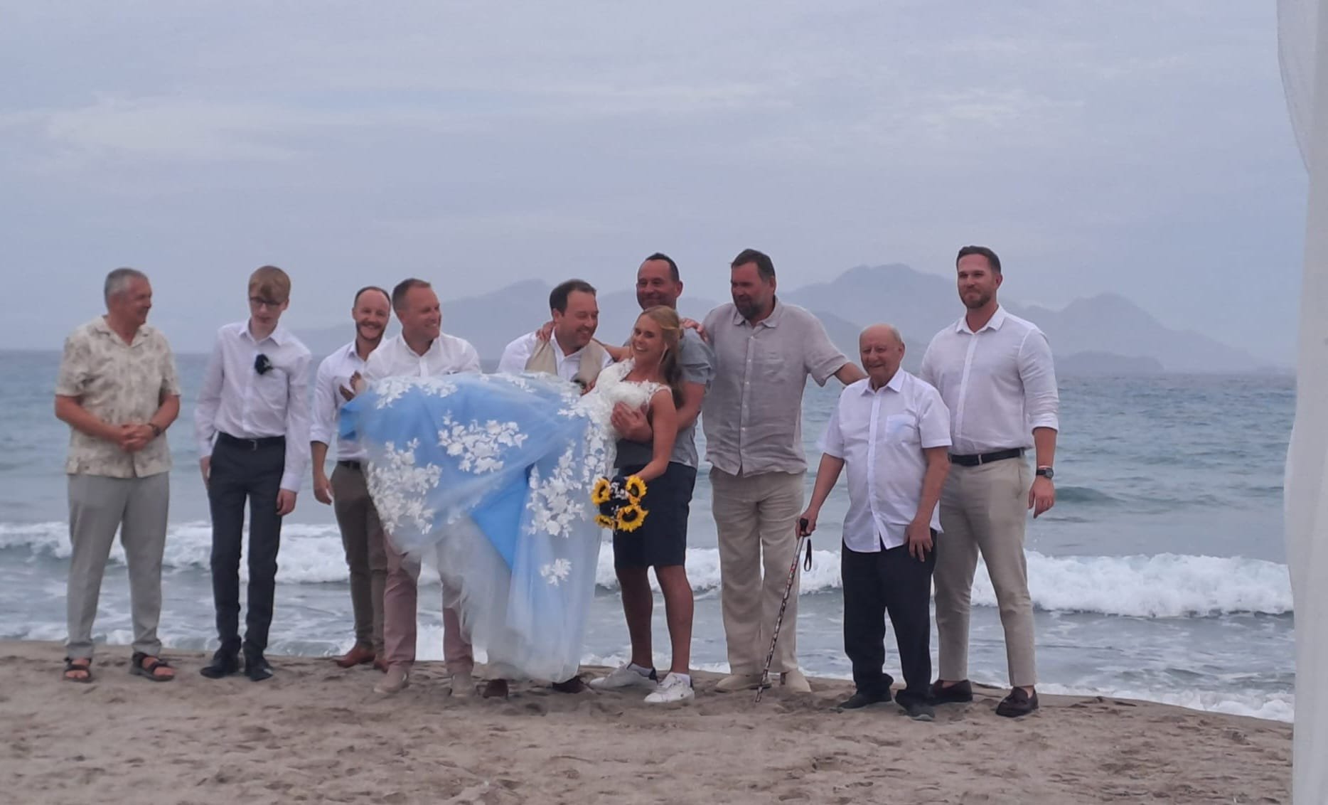 Group of people on a beach, with a bride in a white dress being carried by a man, surrounded by friends and family, during a wedding ceremony near the ocean with mountains in the background.