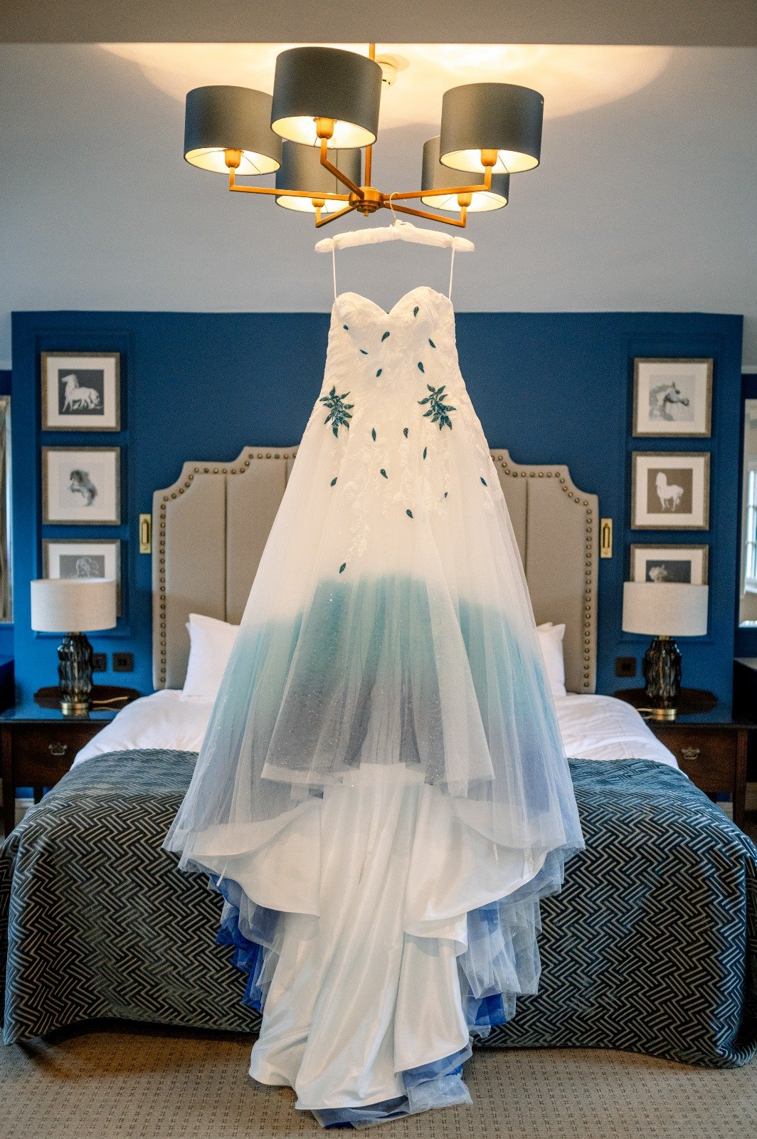 A wedding dress with floral and leaf embroidery hanging in a bedroom with blue walls, framed horse pictures, and bedside lamps.