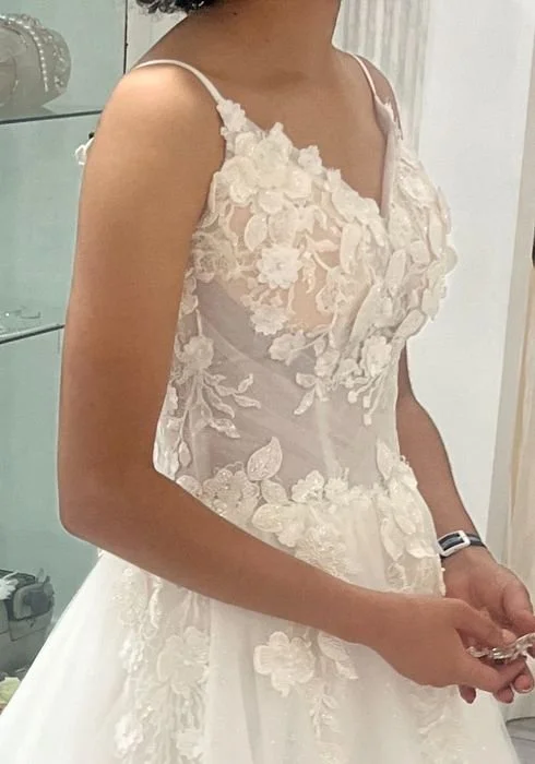 Close-up of a woman wearing a white wedding dress with floral lace details on the bodice and straps, standing indoors.