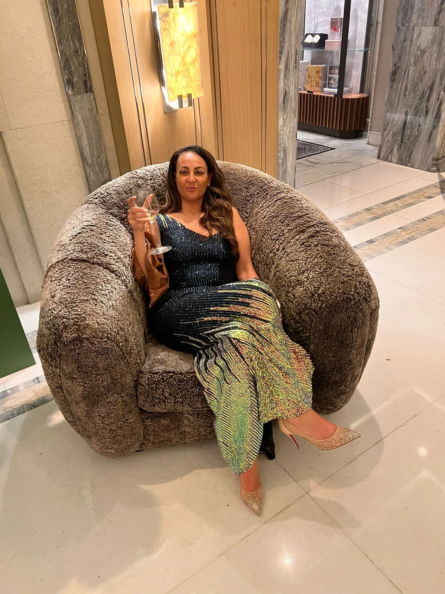 A woman in a sparkly black and gold dress sitting on a plush, round, brown chair, holding a glass of white wine in her right hand, wearing beige high heels with small embellishments, in a hotel lobby with marble walls and floor, and a display case in