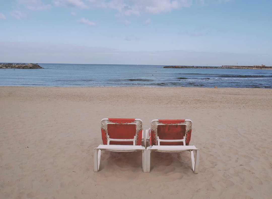 Two empty red and white beach lounge chairs on sandy beach facing the ocean with a pier or breakwater in the distance under a partly cloudy sky.