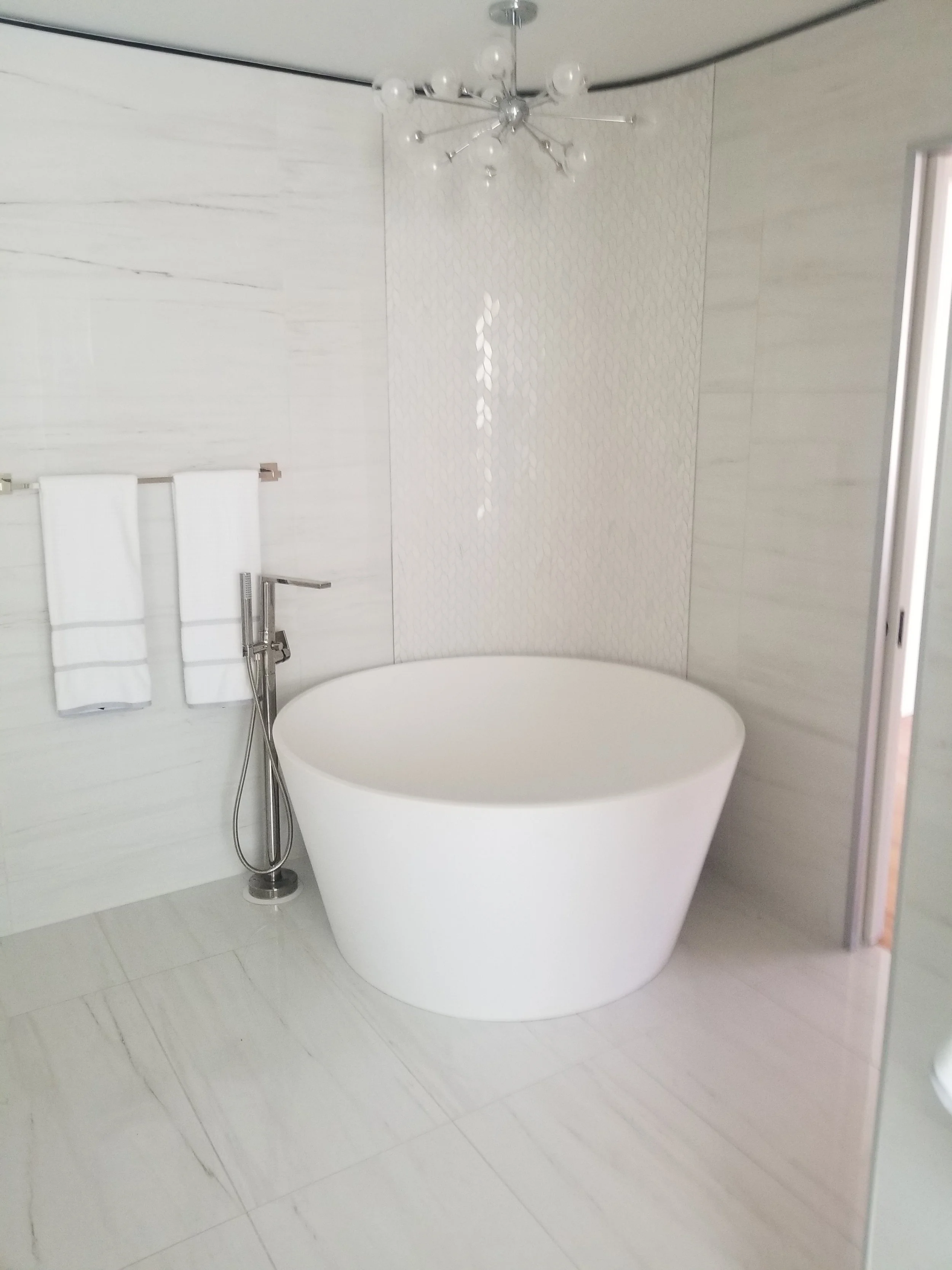 Modern bathroom with a white freestanding bathtub, wall-mounted towel rack with two white towels, handheld showerhead, and a unique starburst chandelier.