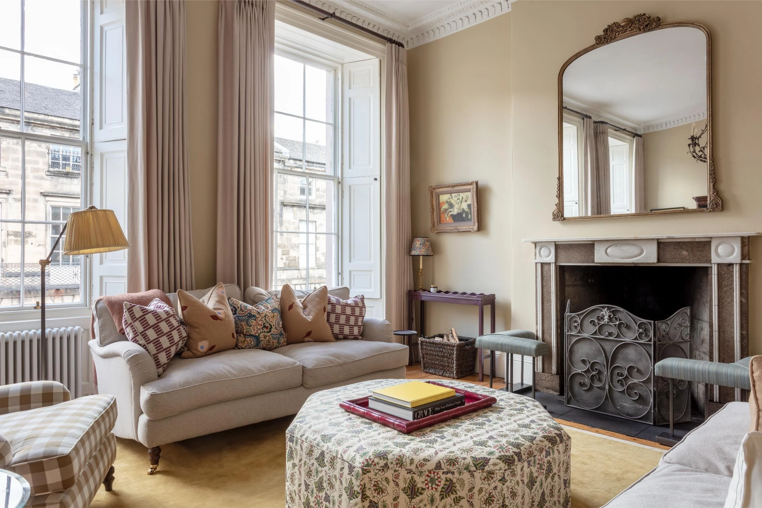 A living room with large windows, beige walls, a fireplace with a mirror above it, and various furniture including a couch, armchair, coffee table with books, and side tables with lamps.