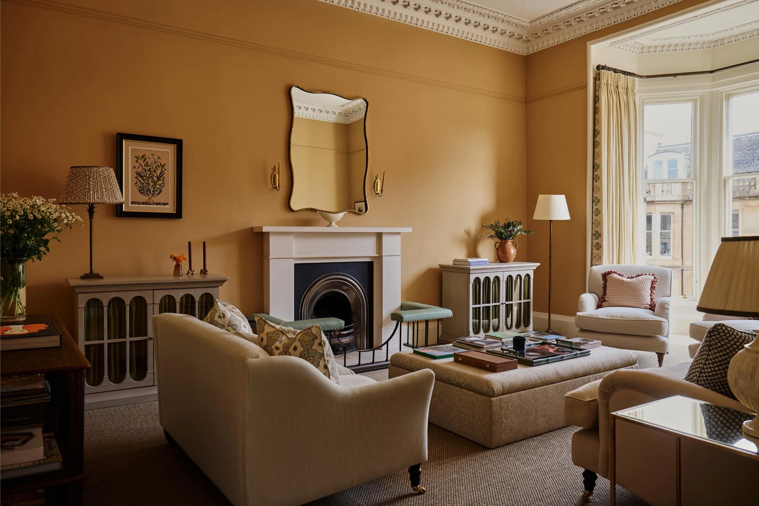 Living room with yellow walls, cream furniture, fireplace, mirror, plants, framed artwork, and large window with curtains
