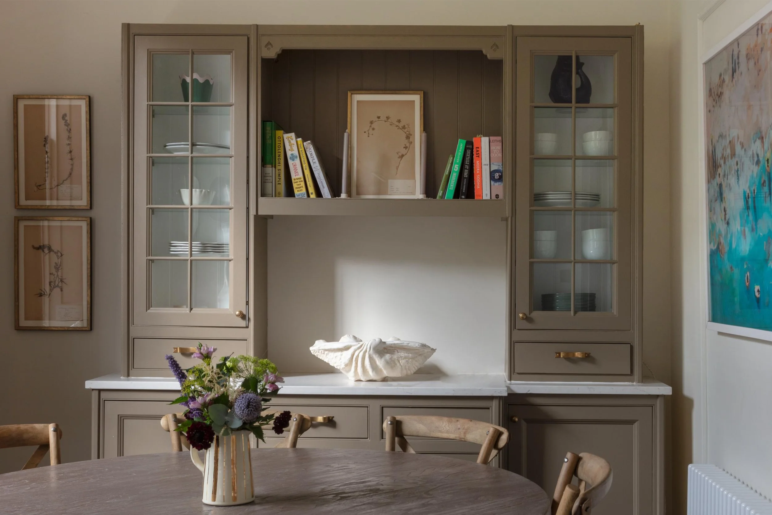 Dining room with a round wooden table, surrounded by chairs, and a decorative vase with purple flowers on top. Wall-mounted cabinet with glass-paneled doors, open shelves with books and ceramics, framed botanical prints on the wall, a large colorful 