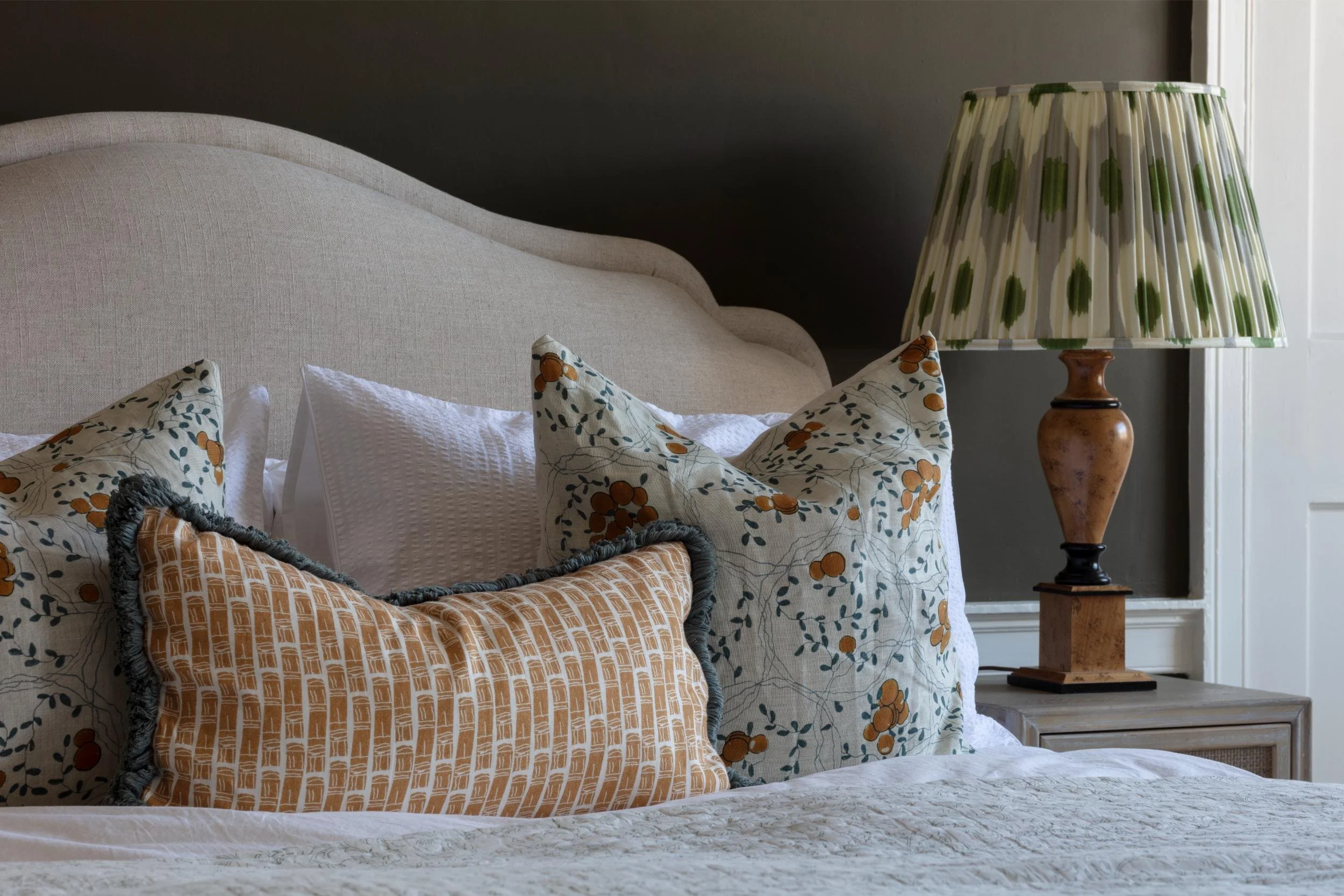 A bedroom scene featuring a white cushioned headboard, decorative pillows with floral and geometric patterns, a beige and brown wicker basket, a wooden bedside table, and a vintage-style table lamp with a green and white patterned lampshade.