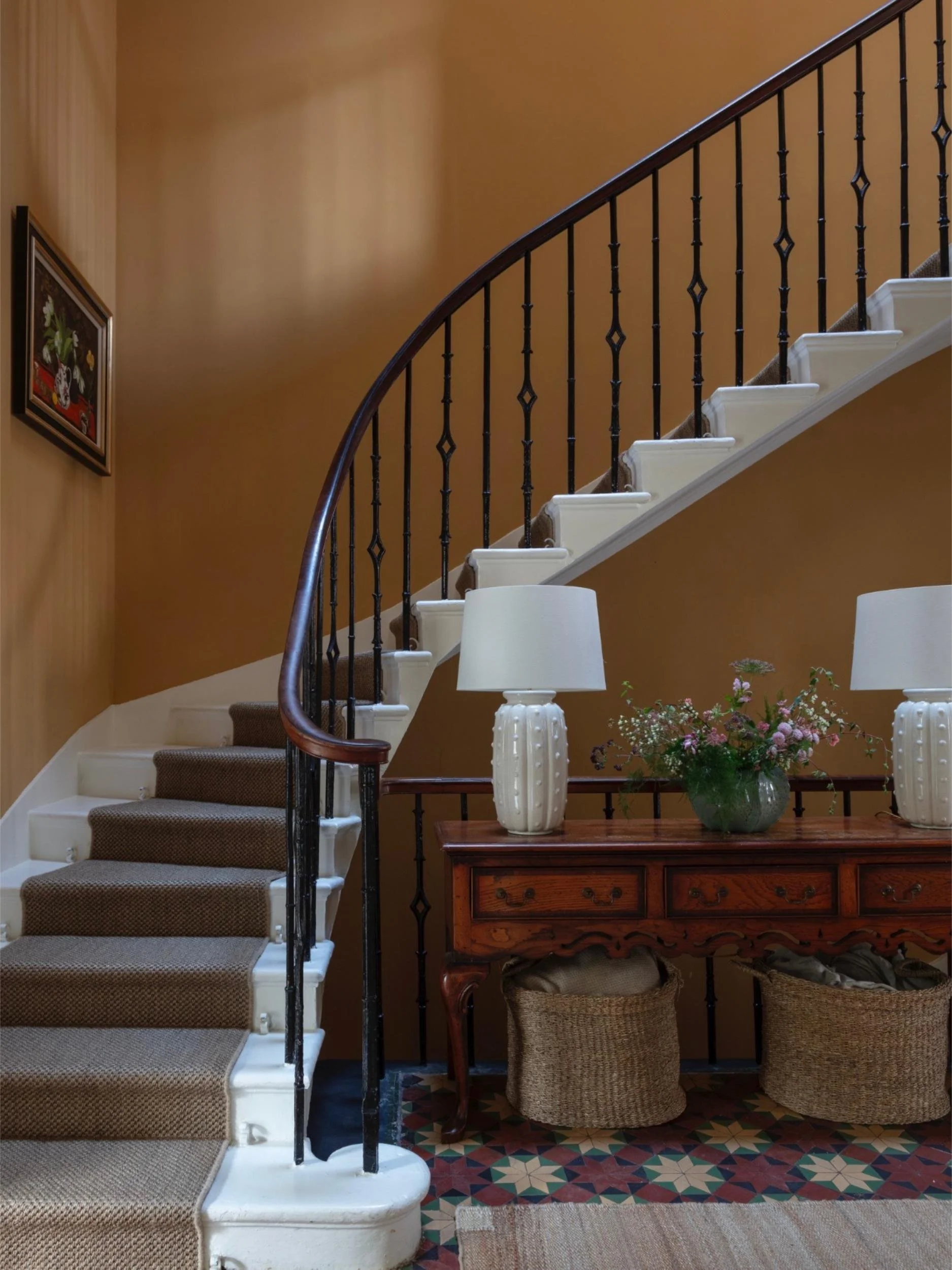 Staircase with carpeted steps and a wooden handrail, beside a wooden console table with vases and a flower arrangement, against a beige wall with a framed painting.