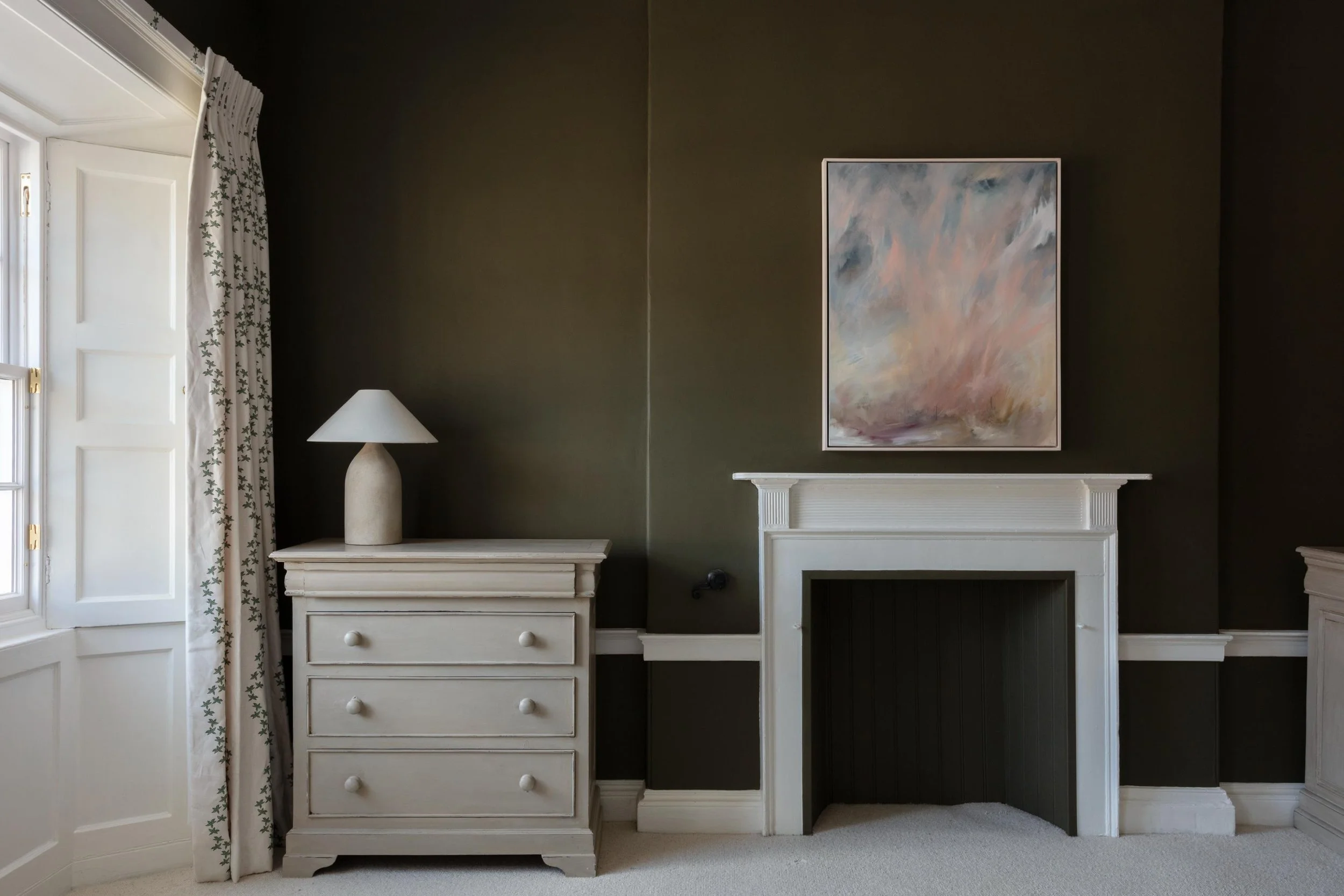 A living room corner with a dark wall, white fireplace mantel, a framed abstract painting above it, a white dresser with five drawers and round knobs, topped with a large cream-colored lamp, and a window with patterned curtains.