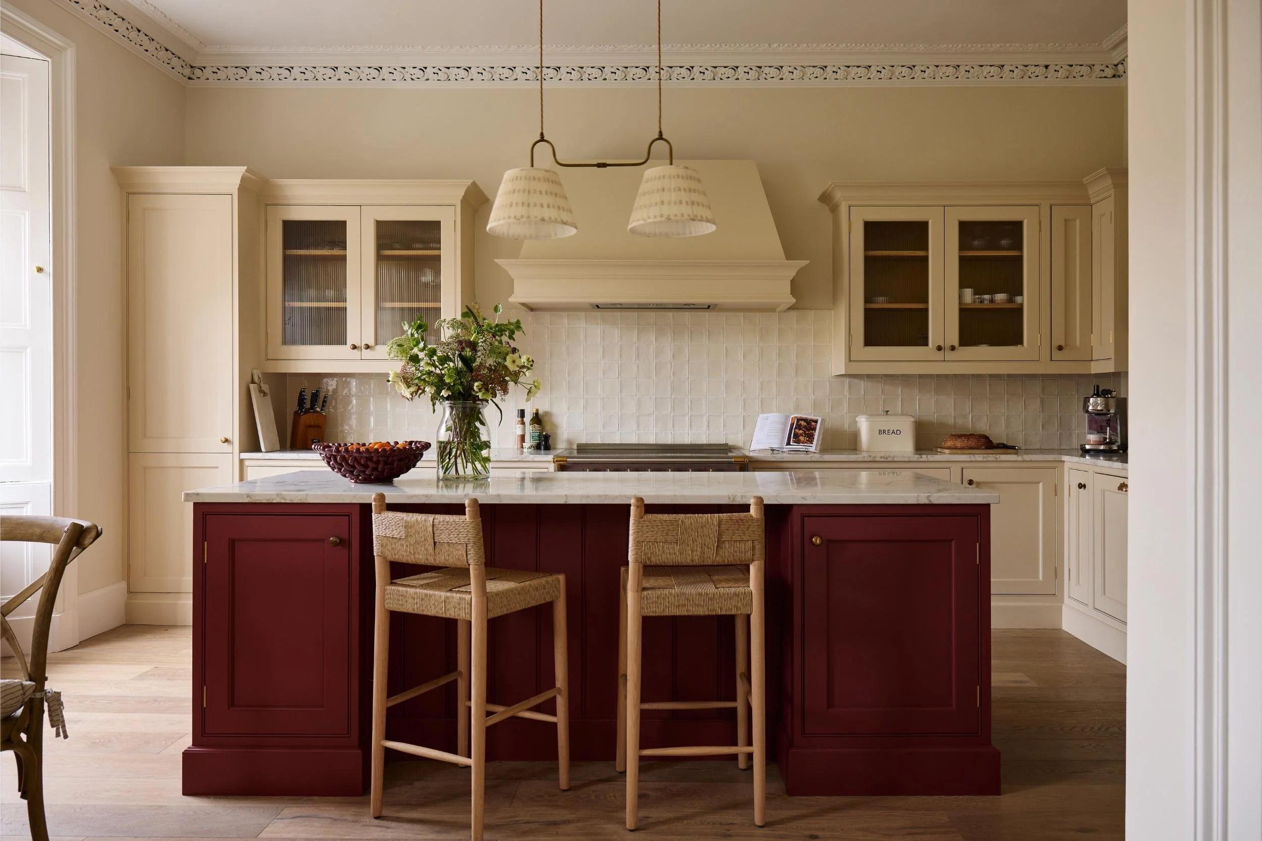 A kitchen with beige cabinets, a red island, a white marble countertop, hanging light fixtures, a vase of flowers, a fruit bowl, and a cookbook. Other kitchen appliances and items are visible on the counter.