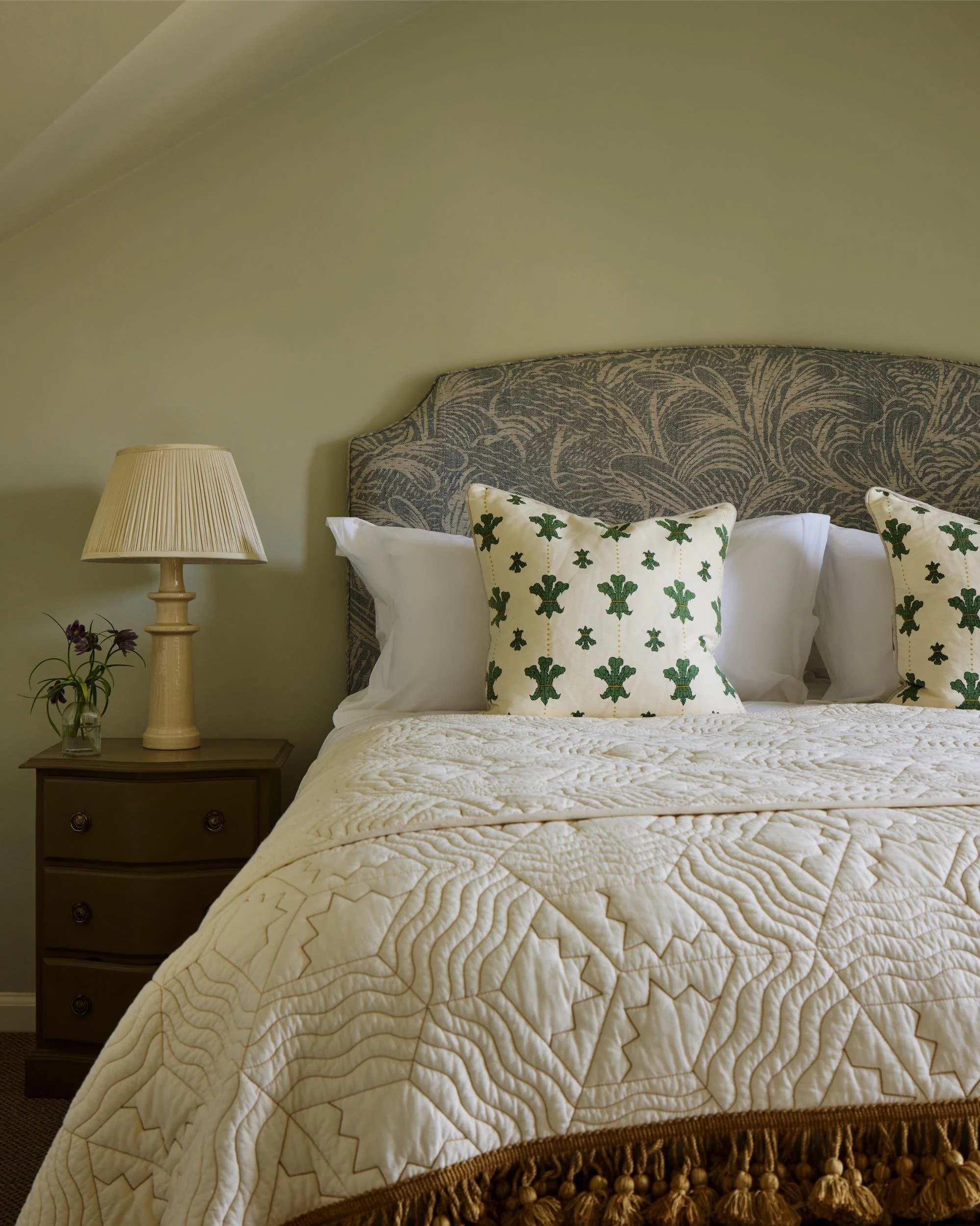 A neatly made bed with white quilted bedding, decorative pillows with green shamrocks, a gray patterned headboard, a nightstand with a beige lamp, and a small vase of purple flowers against a pale green wall.
