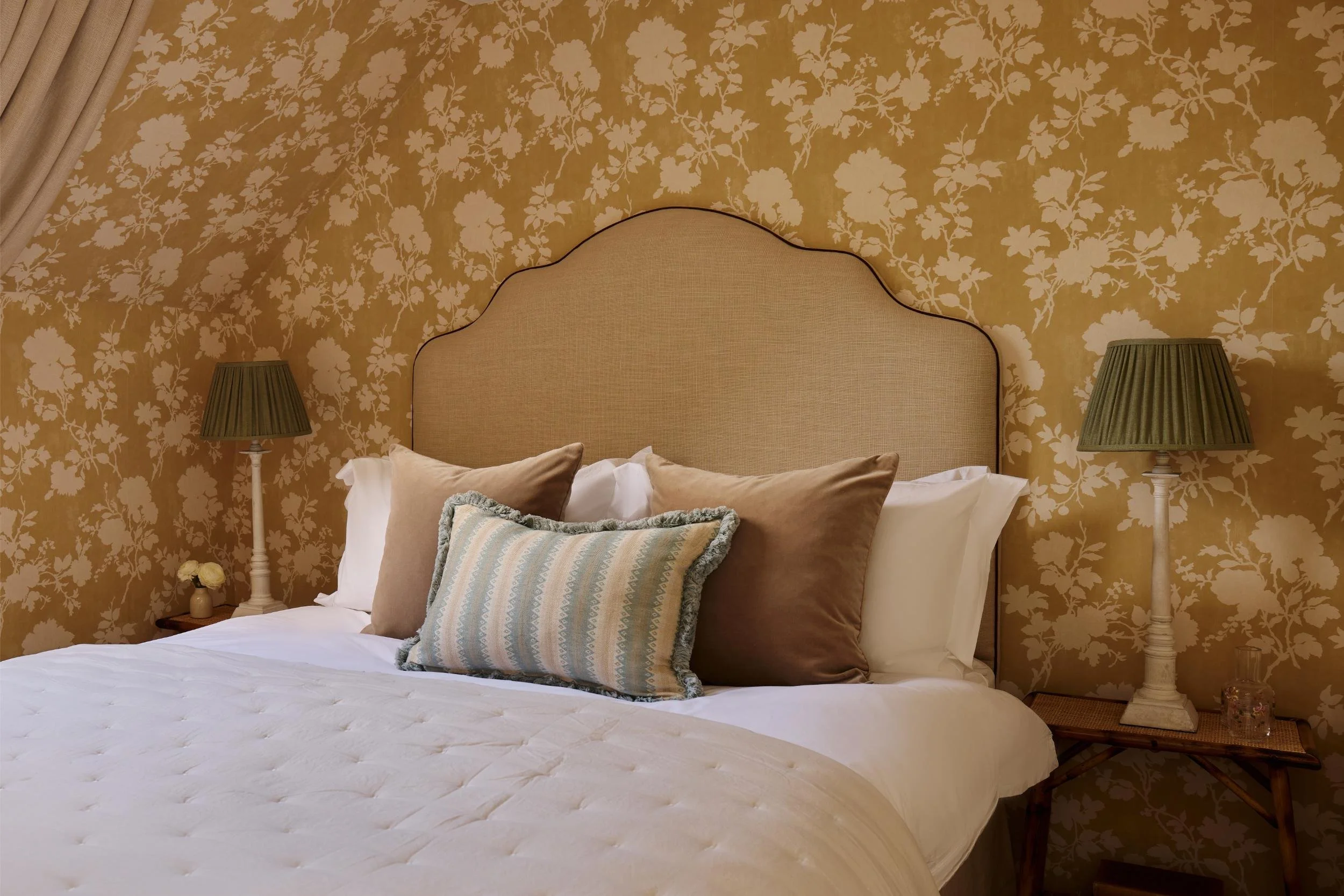 A cozy bedroom with a beige upholstered headboard, patterned wallpaper in yellow with white floral design, two matching green table lamps, and a variety of pillows on the bed.